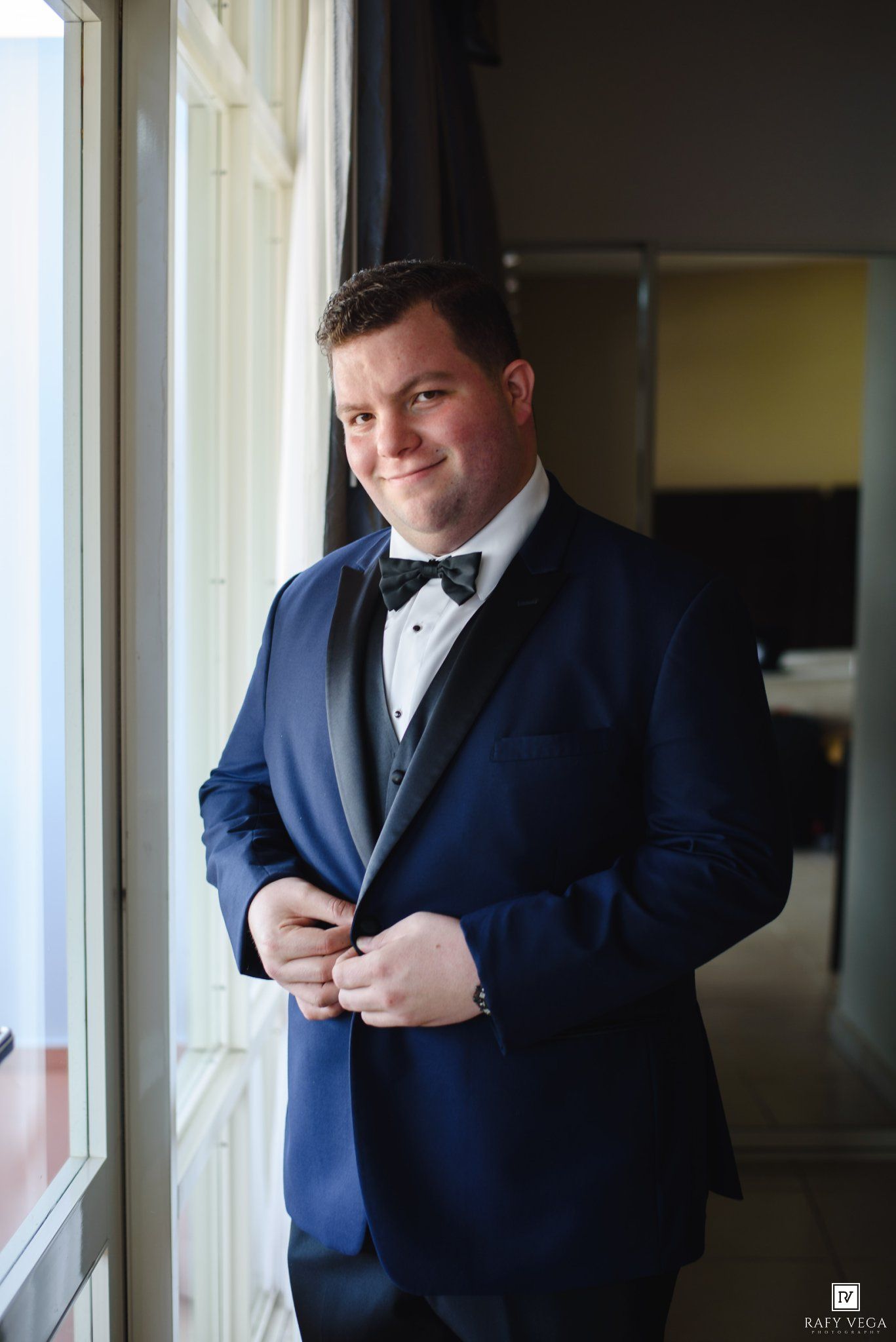 Groom in navy blue tuxedo with black bow tie buttoning jacket while standing near window in hotel room.