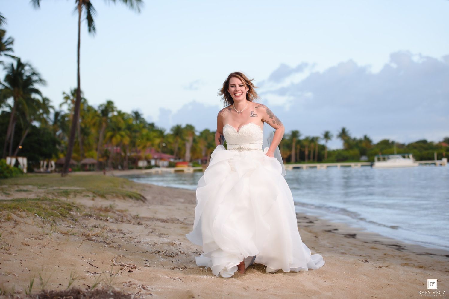 Bride in a strapless white ballgown poses on a tropical beach with palm trees and calm waters at sunset.
