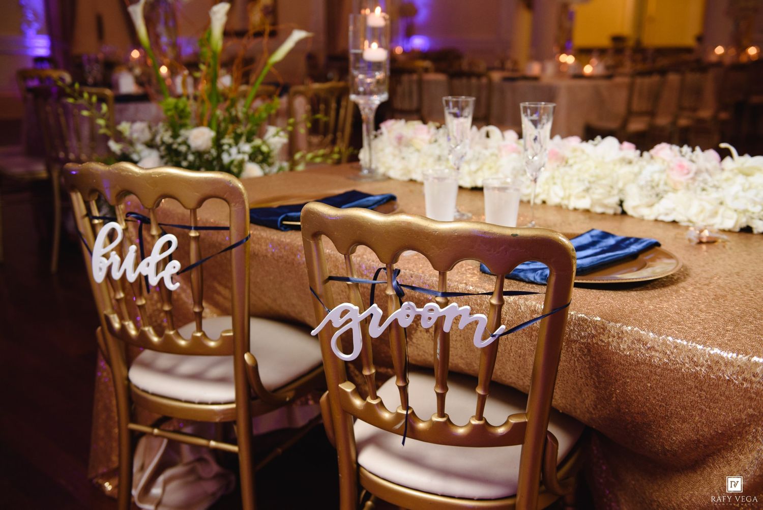 Gold Chiavari chairs with bride and groom signs at an elegant wedding reception table with floral centerpieces.
