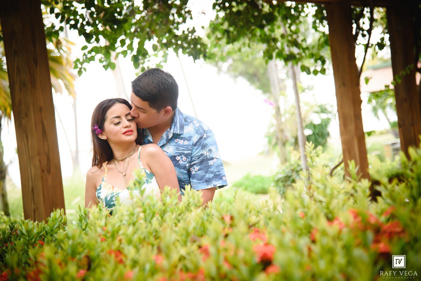 A couple shares a romantic moment among lush green foliage and red flowers, with the man gently kissing the woman's forehead.