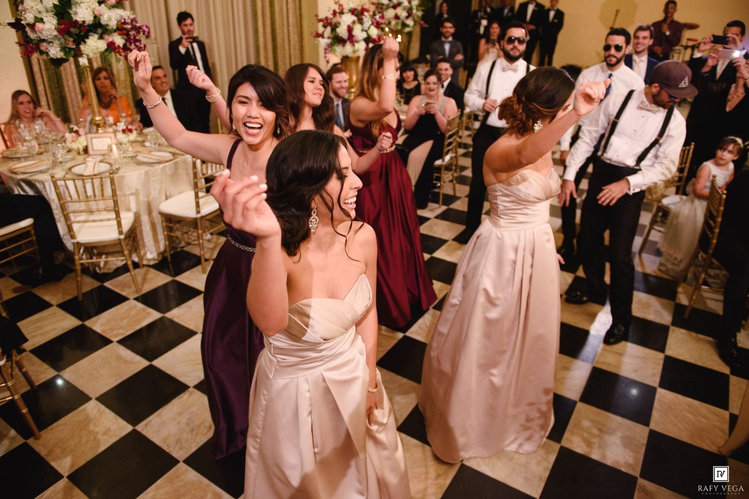 Bridesmaids in blush and burgundy gowns dancing joyfully on a black and white checkered floor at an elegant wedding reception.