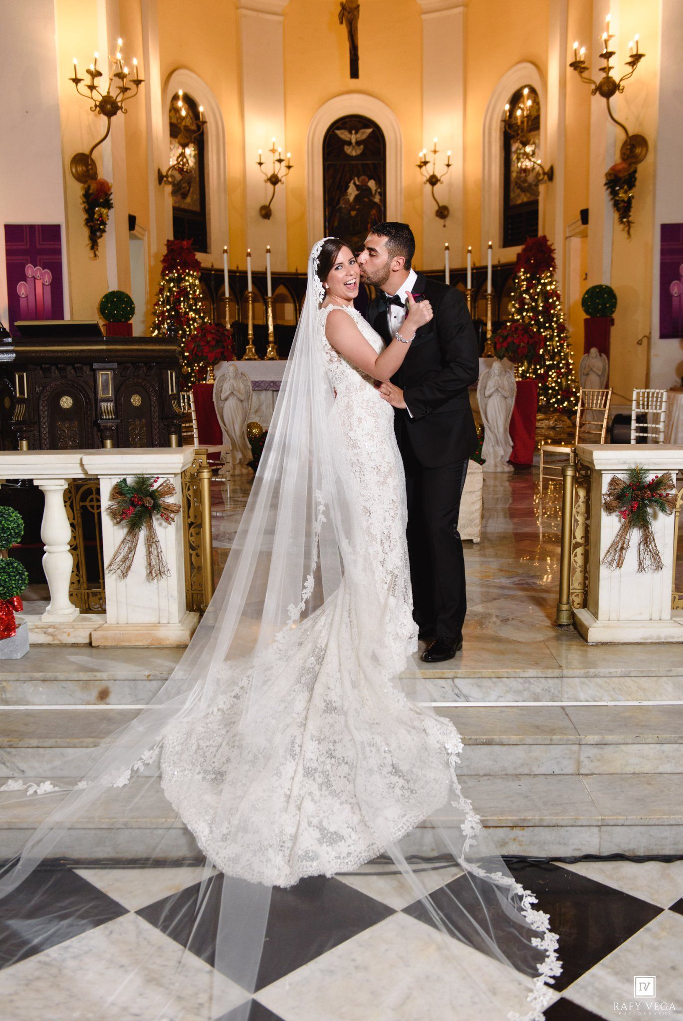 Bride and groom share a romantic kiss at the altar of an elegantly decorated church with Christmas trees and candles.