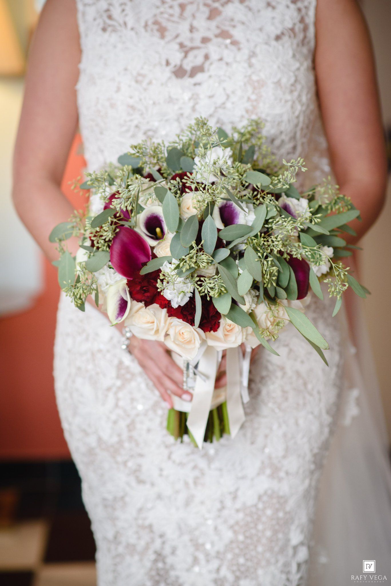 Bride in lace wedding gown holding lush bouquet of burgundy calla lilies, cream roses, and eucalyptus greenery.