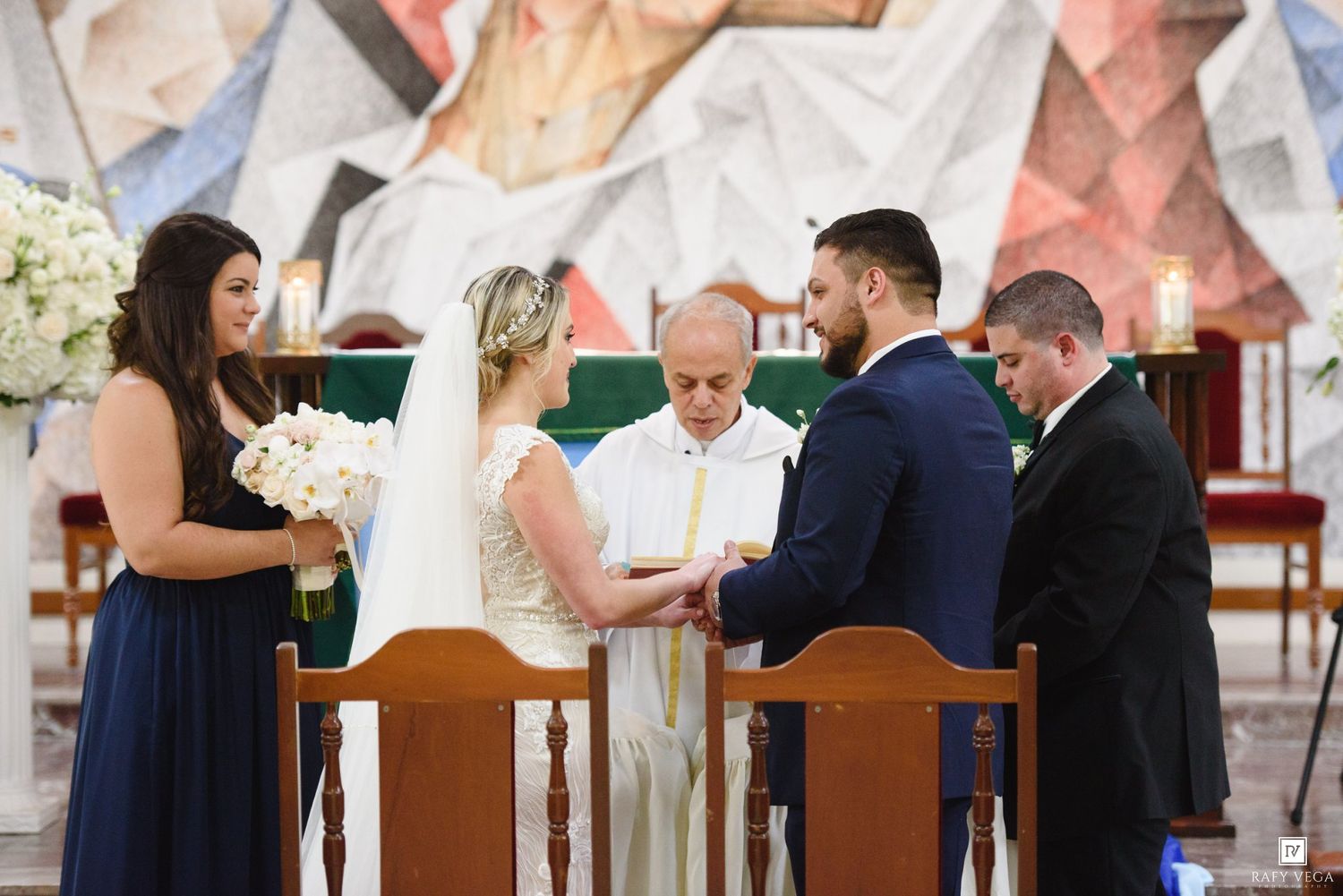 Bride and groom exchange vows at church altar with officiant, bridesmaid, and groomsman during wedding ceremony.