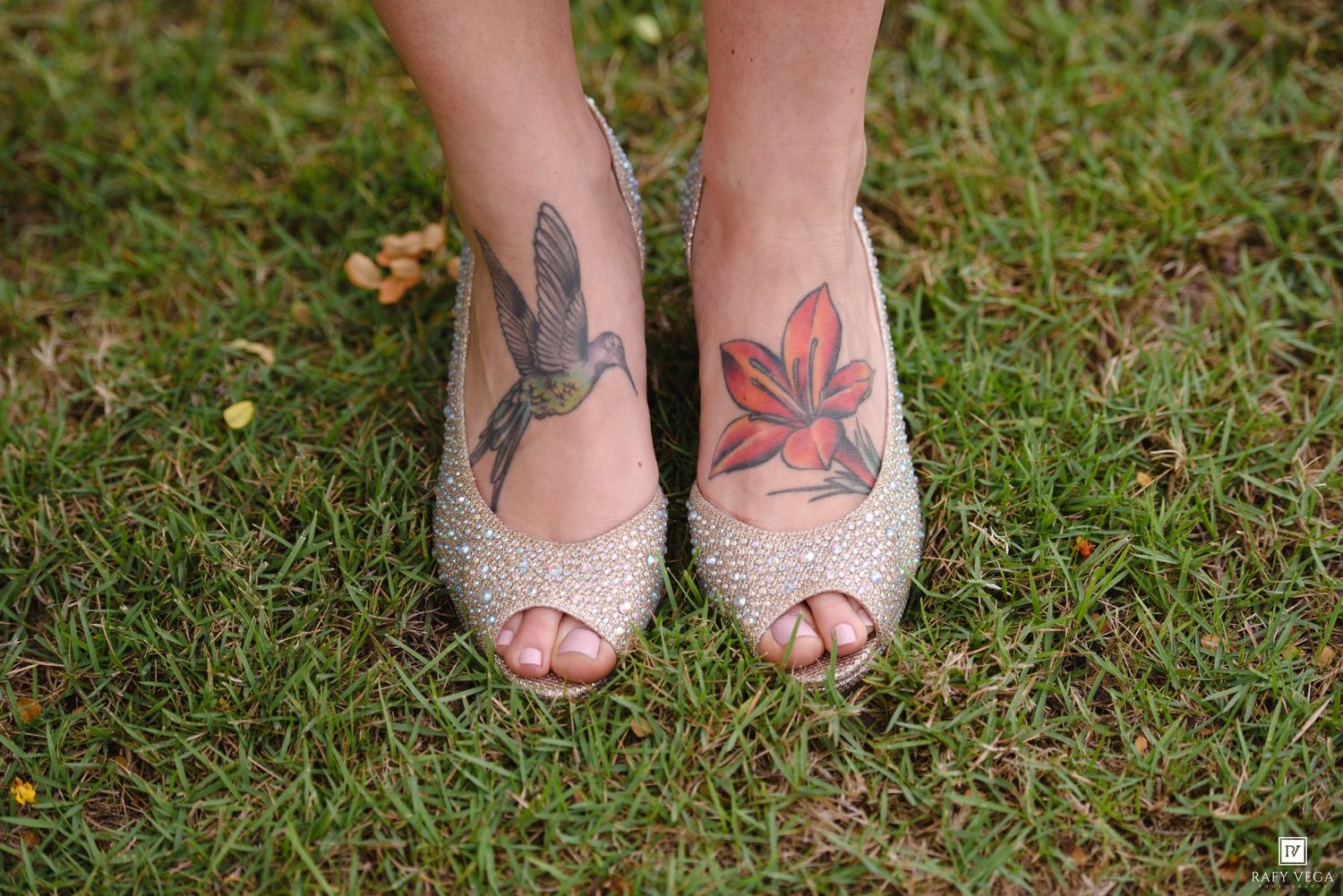 Close-up of bride's feet in sparkling peep-toe heels with hummingbird and floral tattoos on green grass.