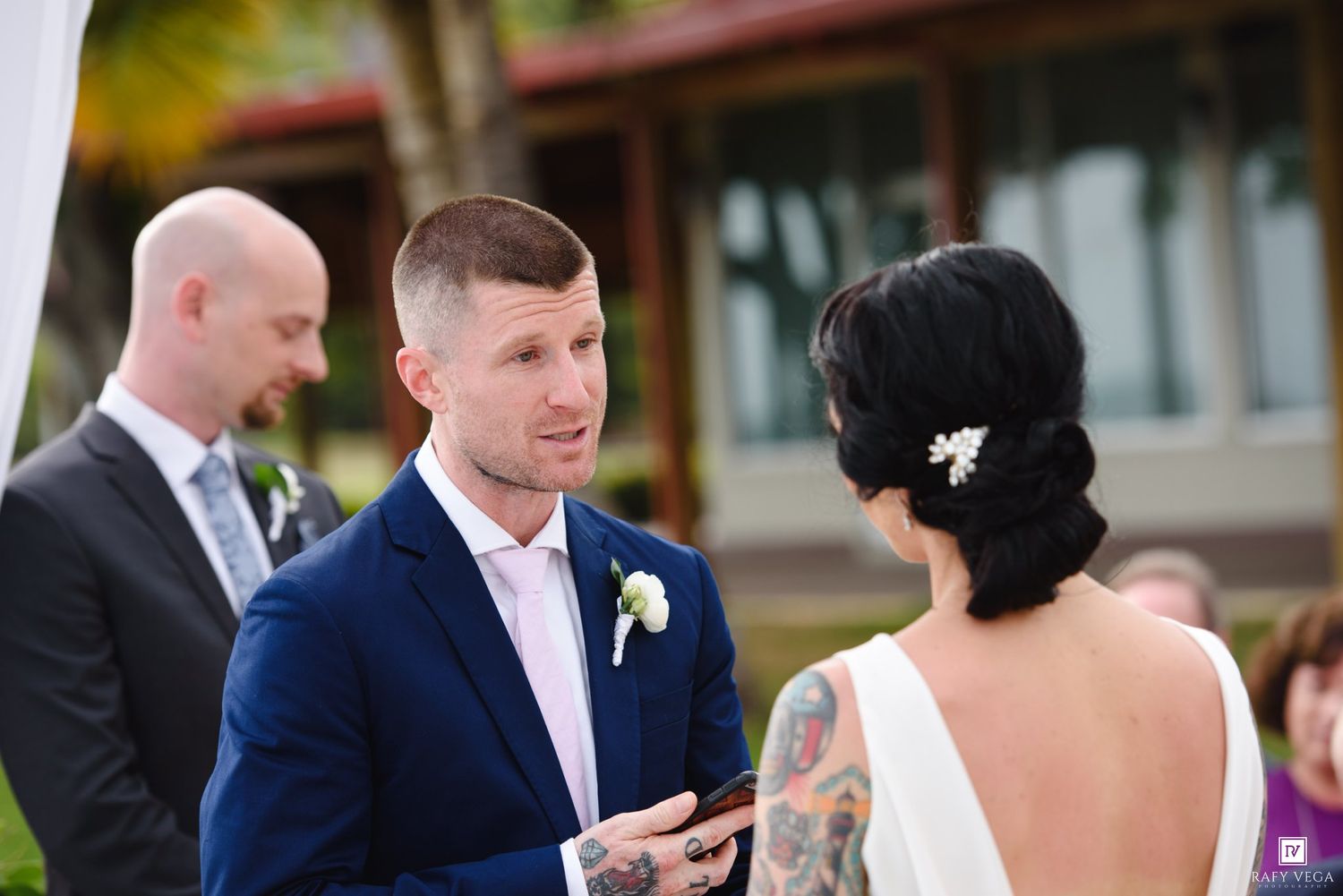 Groom in navy suit reads vows to tattooed bride during outdoor wedding ceremony with guests in background.
