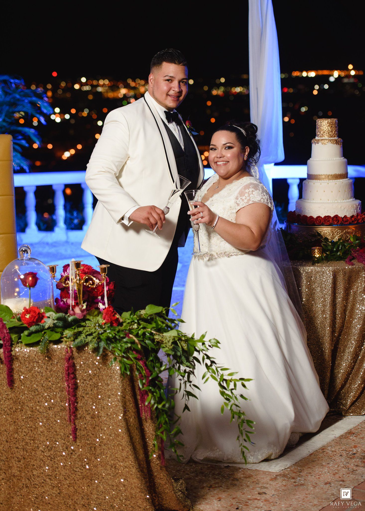 Bride and groom smiling beside wedding cake at elegant outdoor night reception with city lights in background.