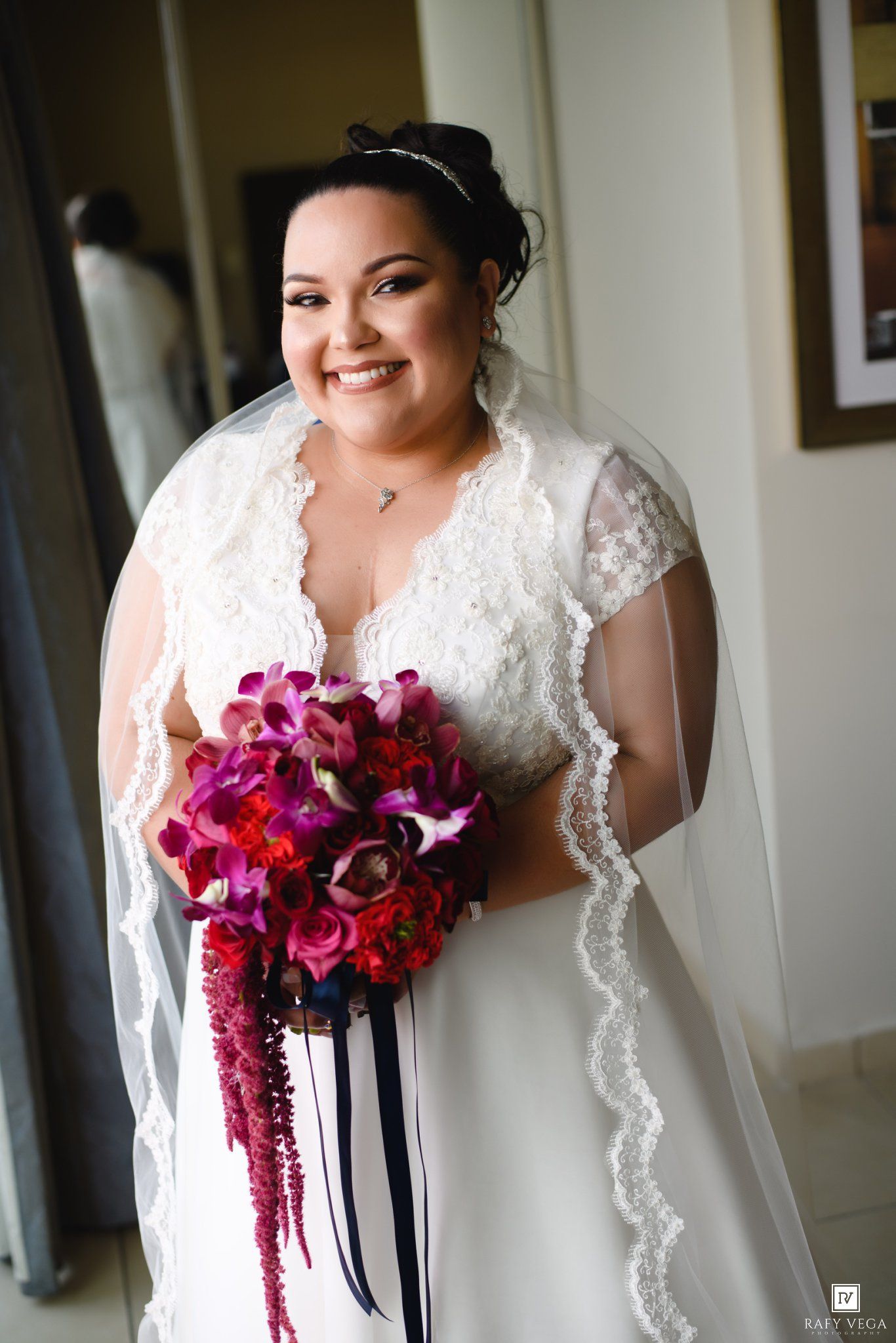 Smiling bride in white lace wedding dress holding vibrant fuchsia and burgundy bouquet with navy ribbon accents.