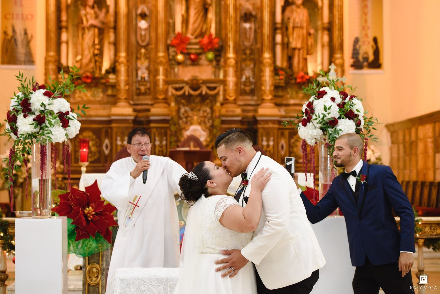 Bride and groom share first kiss at church altar while officiant and guests look on.