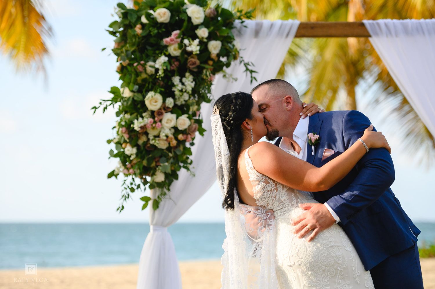 Couple sharing first kiss at beach wedding ceremony under floral arch with palm trees and ocean backdrop.