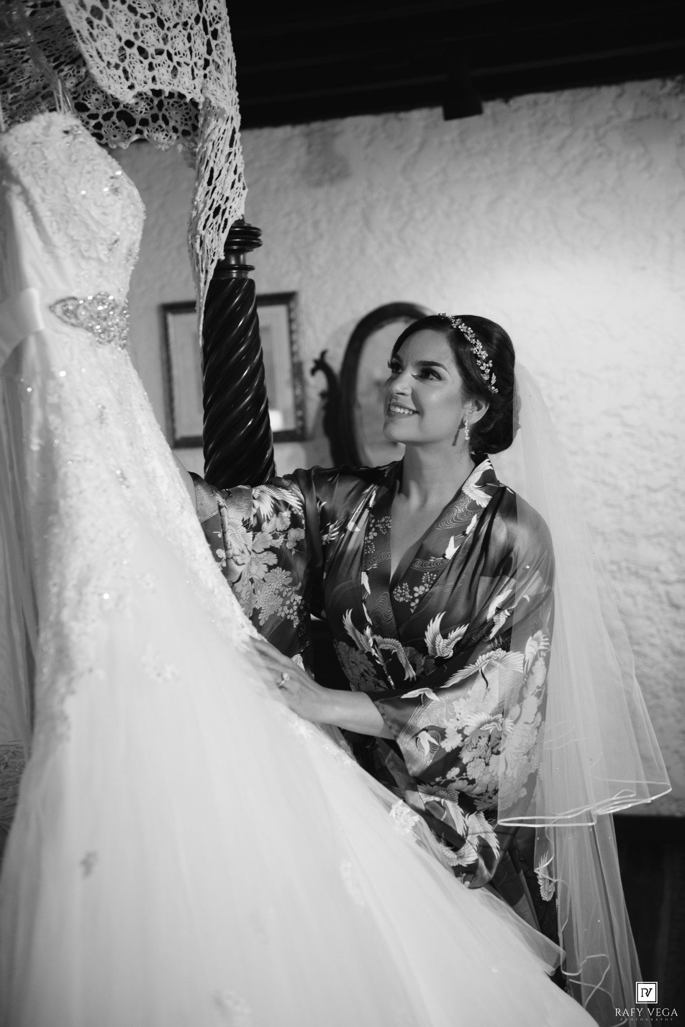 Black and white photo of a bride in a floral robe admiring her wedding dress on a mannequin before the ceremony.