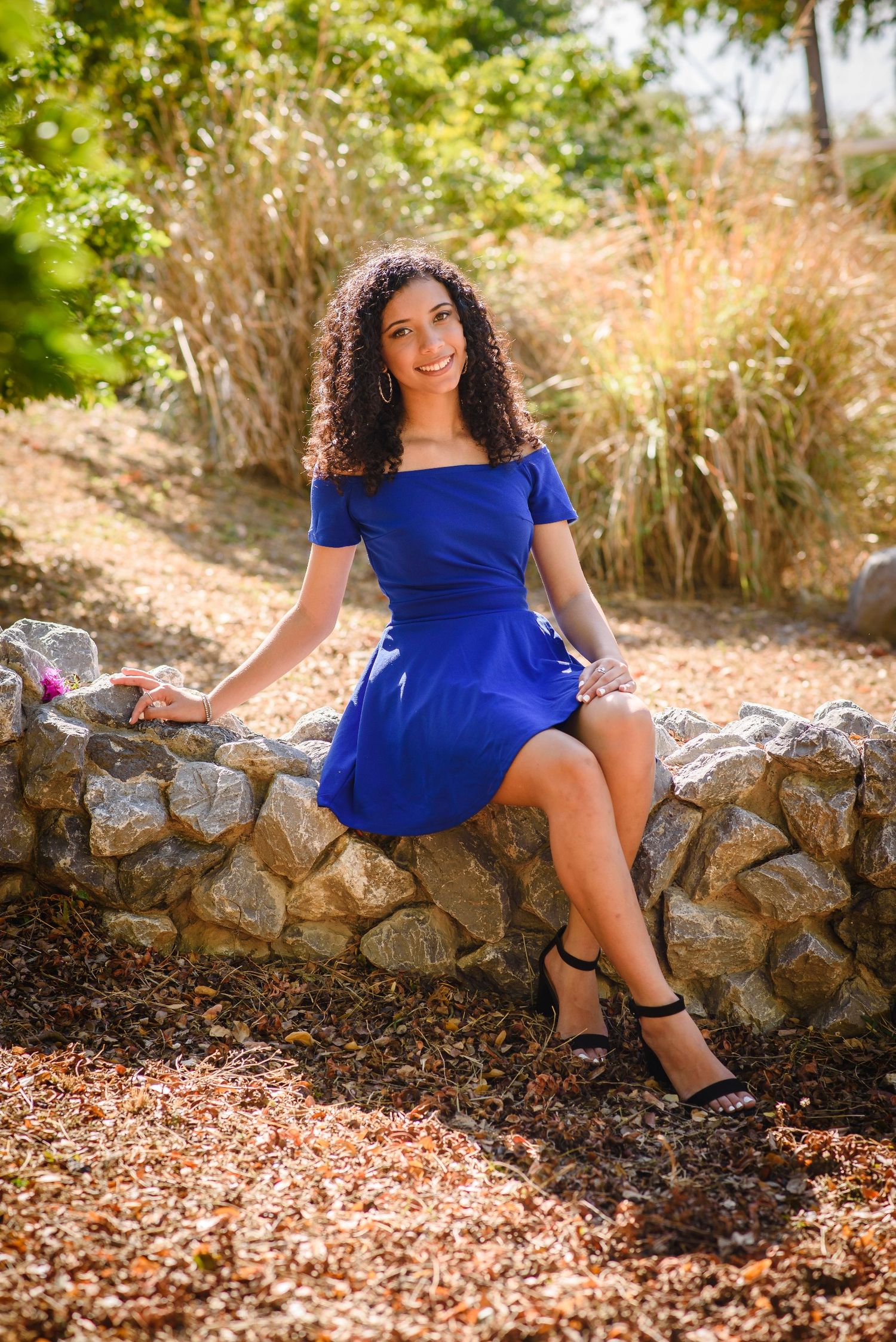 A smiling woman with curly hair sits on a stone wall wearing a blue off-shoulder dress and black heeled sandals outdoors.