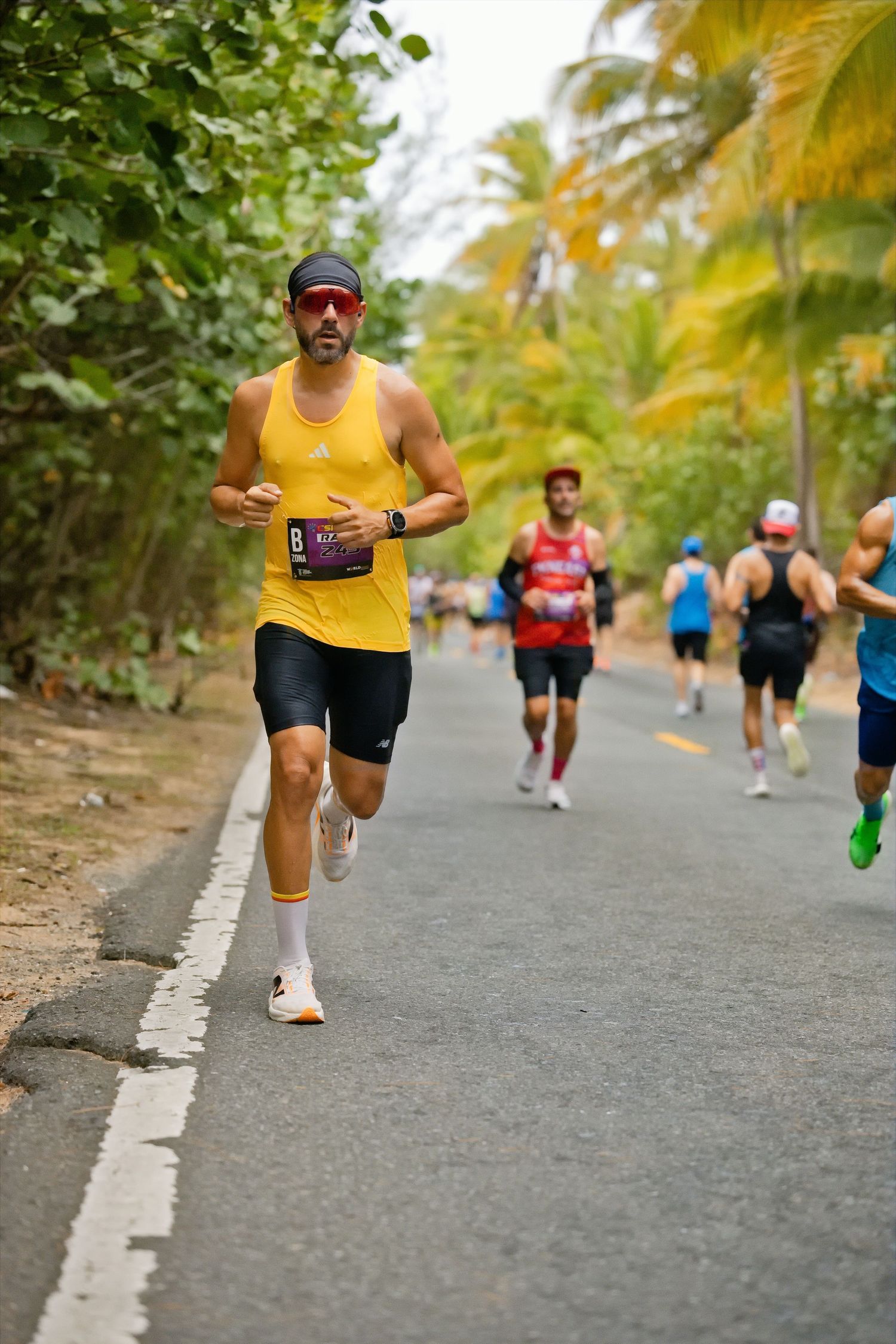 Male runner in yellow vest leads a tropical road marathon race, wearing sunglasses and race bib number 8.