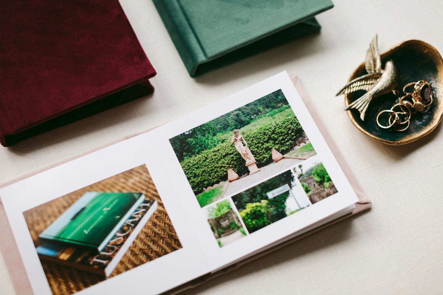 Open photo album displaying wedding photos surrounded by velvet journals and a decorative bird dish with gold rings.