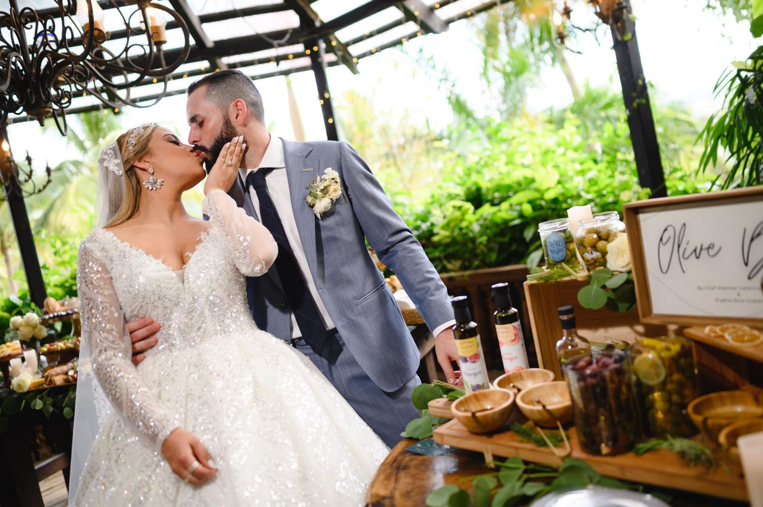 Bride and groom sharing a kiss at their wedding reception near an olive oil tasting table surrounded by lush greenery.