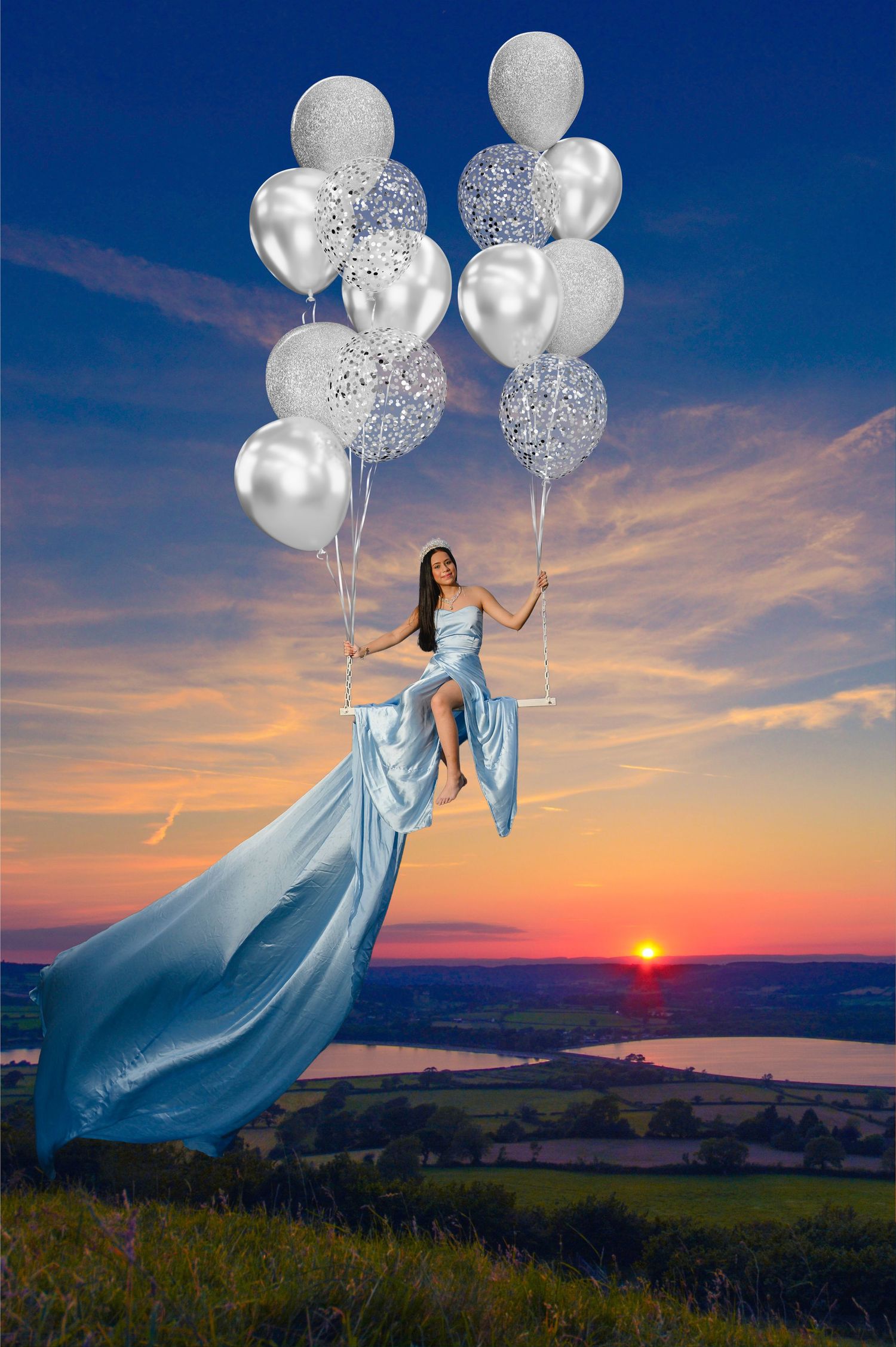 Woman in blue gown sits on swing lifted by silver balloons at sunset over scenic countryside.