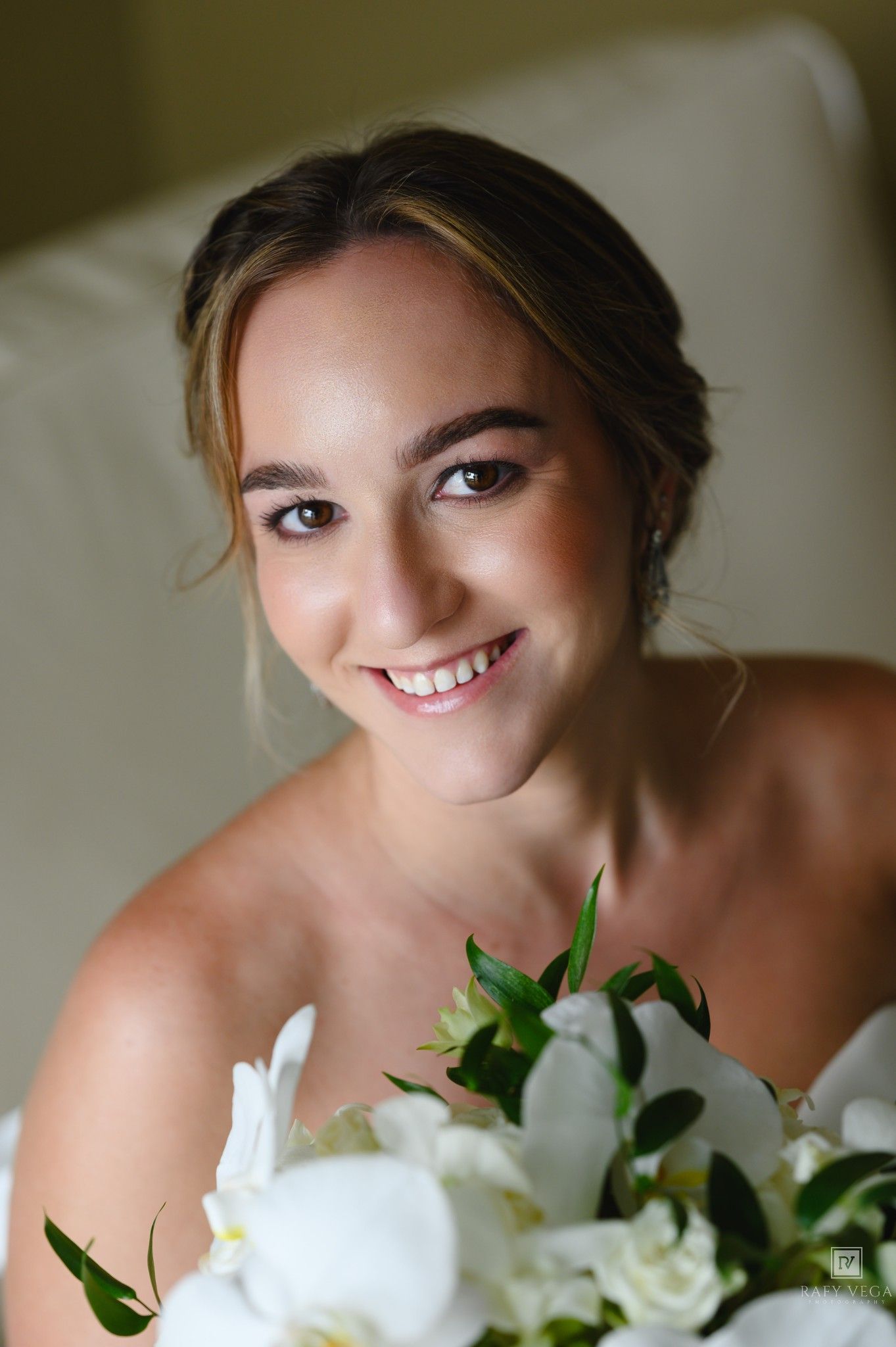 Smiling bride holding white floral bouquet with orchids, wearing strapless gown, natural glam makeup, hair updo.