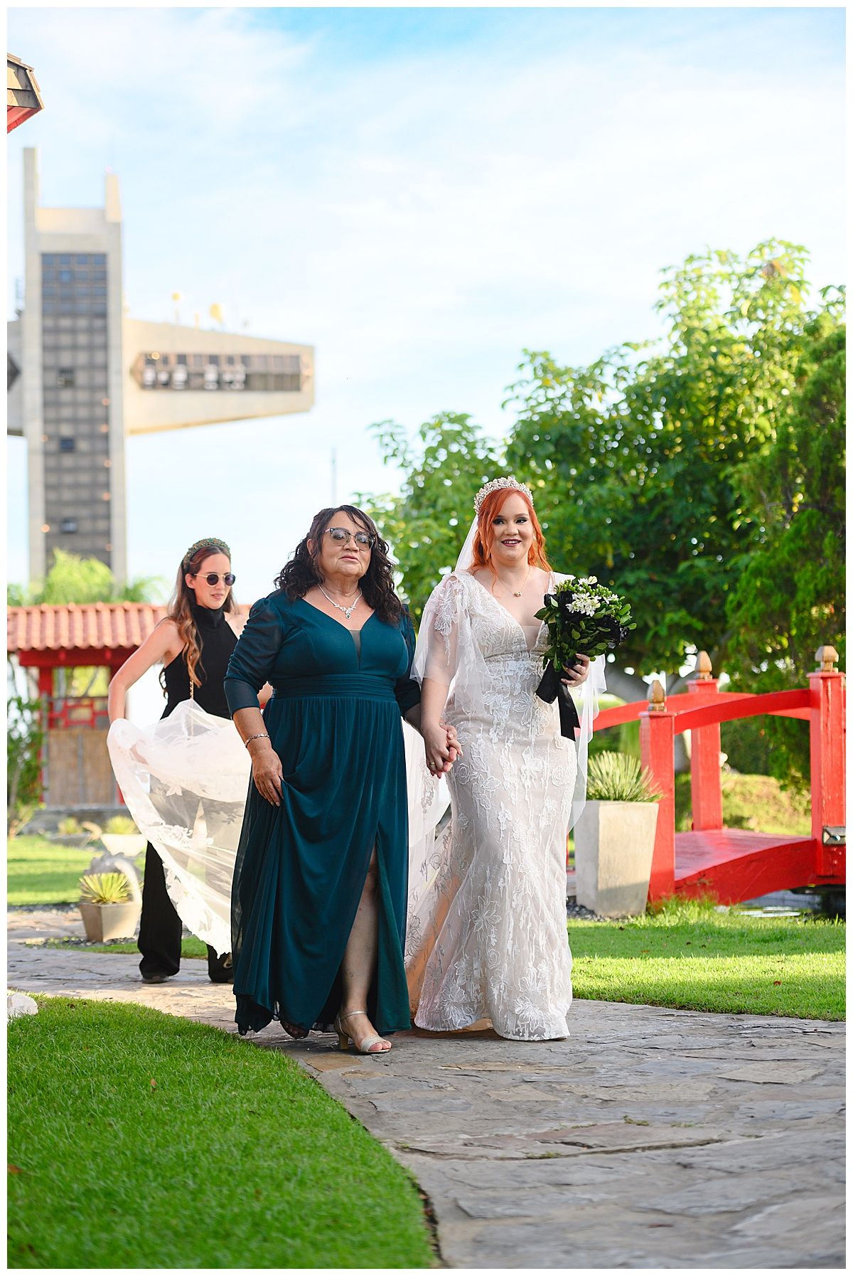 Bride in white lace gown walks with bridesmaids in teal dress along garden stone path.