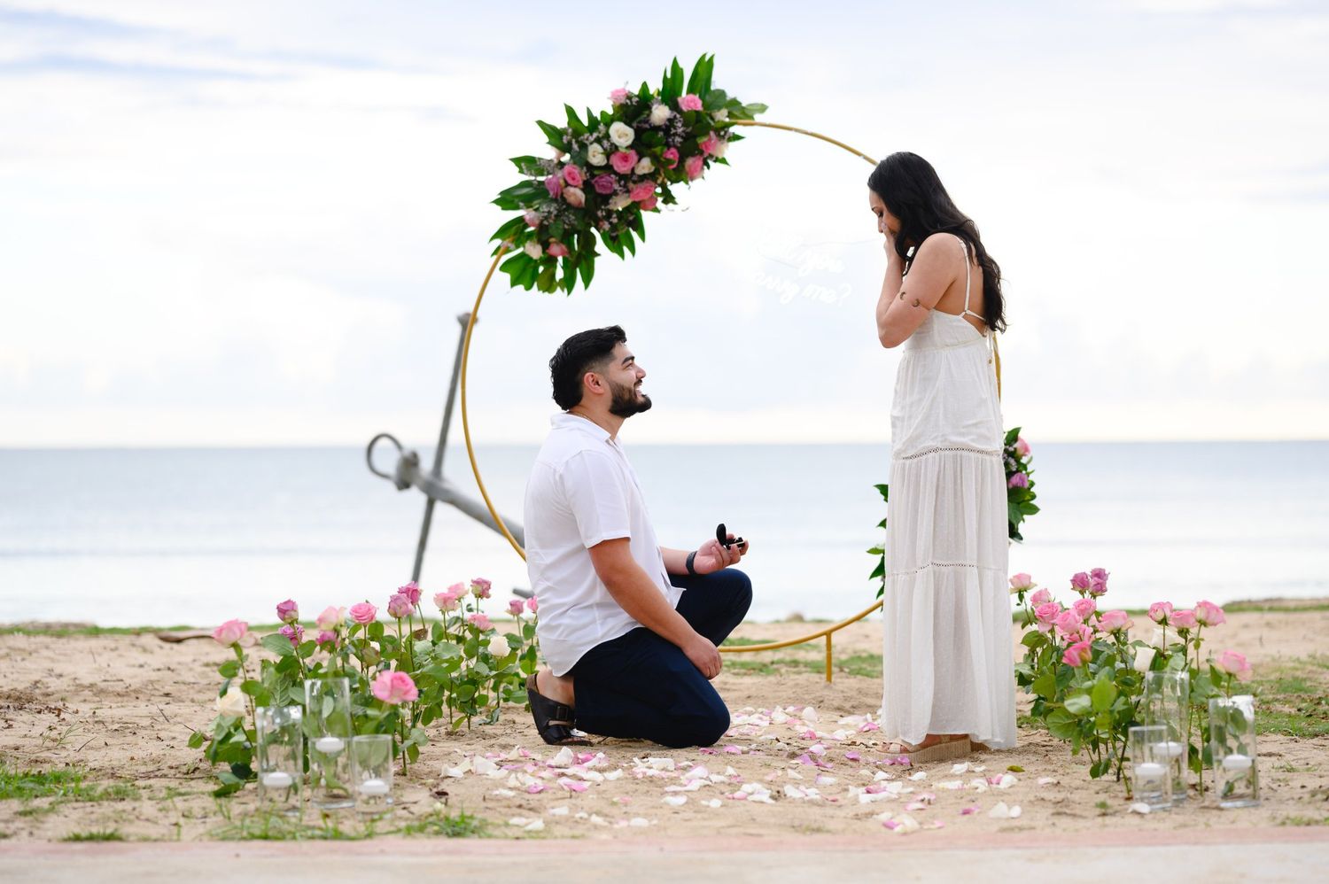 Man proposing to woman on beach under floral arch with pink roses, romantic marriage proposal by the ocean.