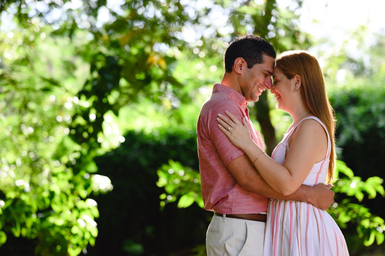Romantic couple embracing face-to-face in a lush green garden, wearing pink and white summer outfits.
