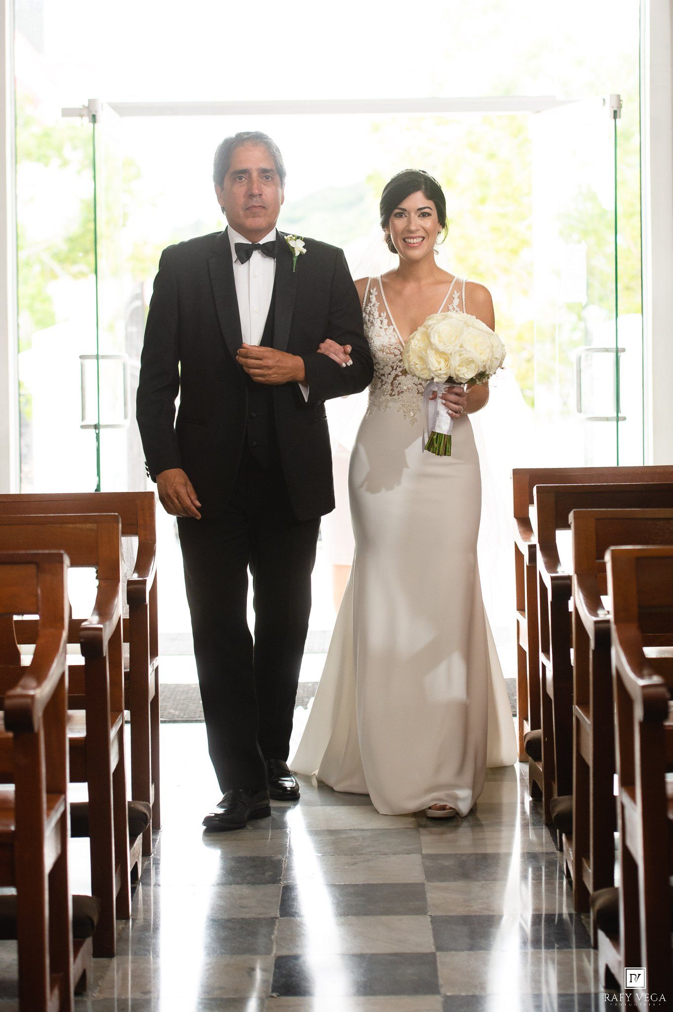 Bride in elegant gown walks down church aisle with escort in tuxedo, holding white bouquet.