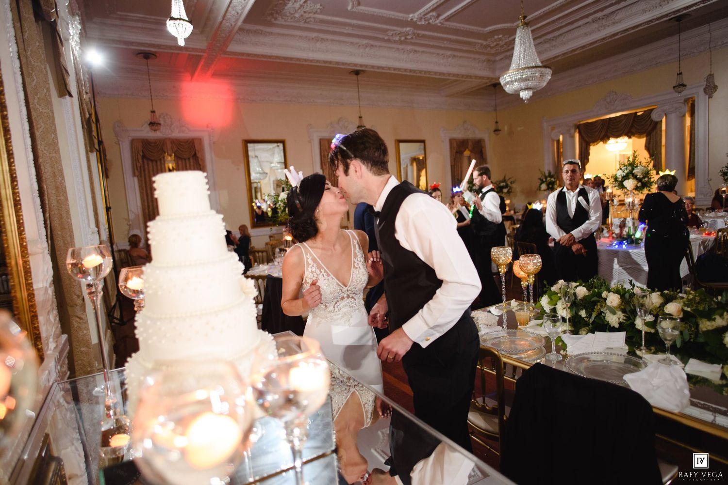 Bride and groom share a romantic kiss beside a white tiered wedding cake at an elegant ballroom reception.