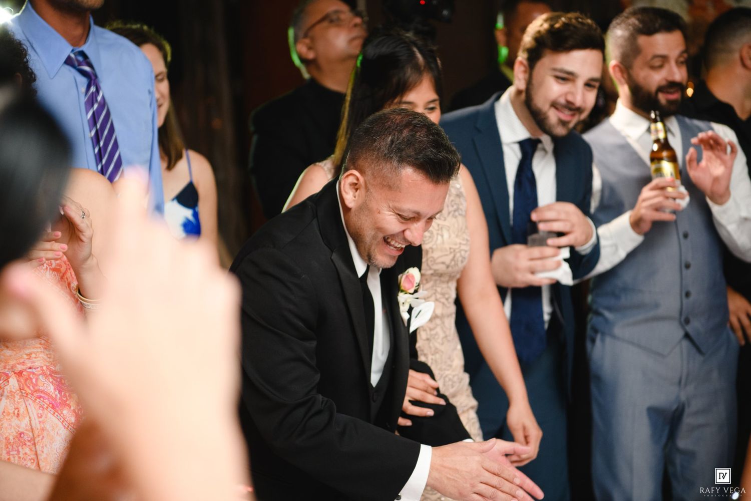 Groom laughing joyfully at wedding reception surrounded by guests celebrating in elegant formal attire.
