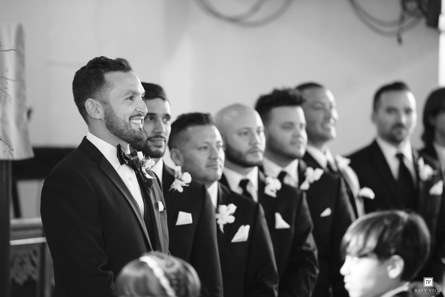 Groom and groomsmen in black tuxedos stand at church altar during wedding ceremony in black and white photo.