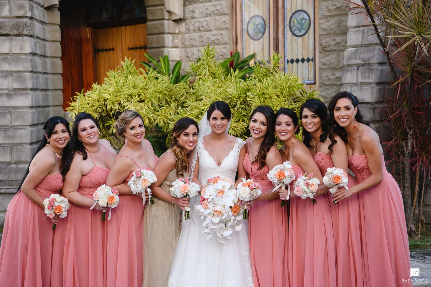 Bride in white gown poses with eight bridesmaids in coral pink dresses holding bouquets outside a stone church.