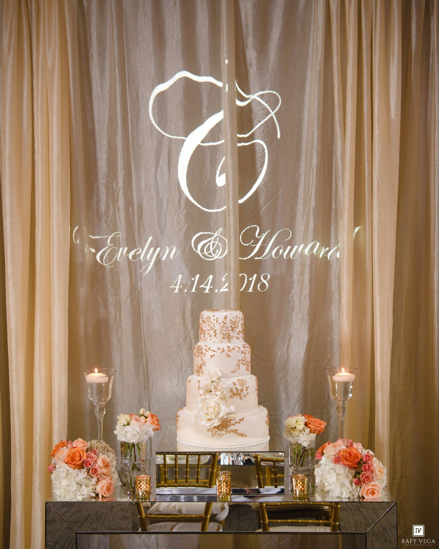 Wedding cake table with monogram backdrop reading Evelyn & Howard 4.14.2018, flanked by candles and peach floral arrangements.