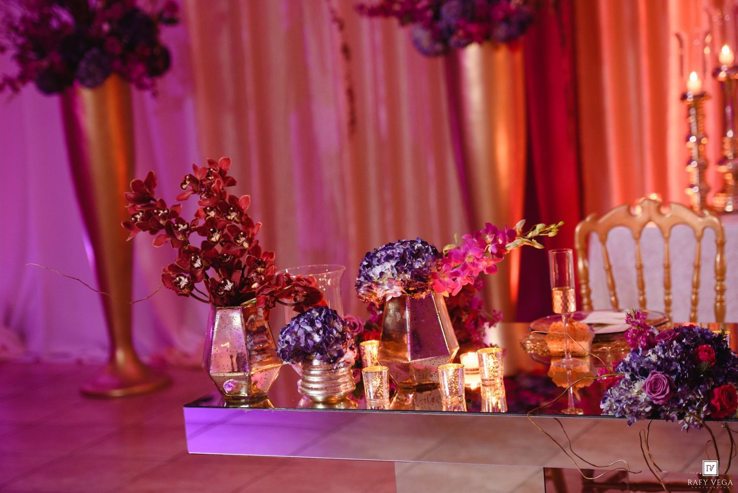 Elegant wedding reception table with purple and red floral arrangements, gold chairs, and glowing candles in a pink-lit ballroom.