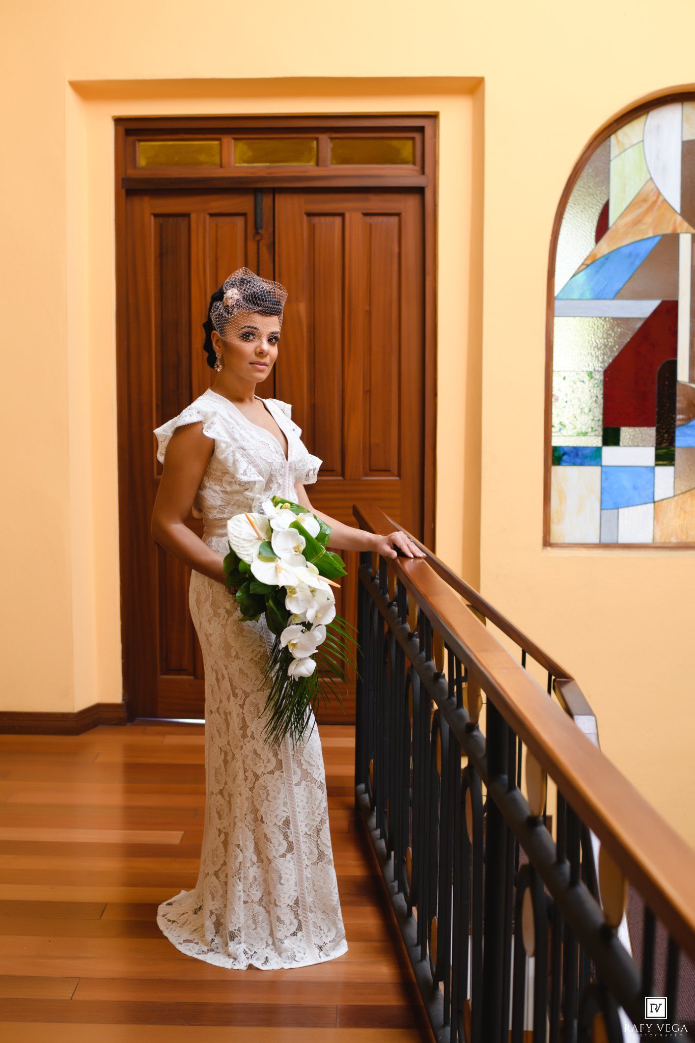 Elegant bride in lace gown holding white calla lily bouquet stands by railing near stained glass window.