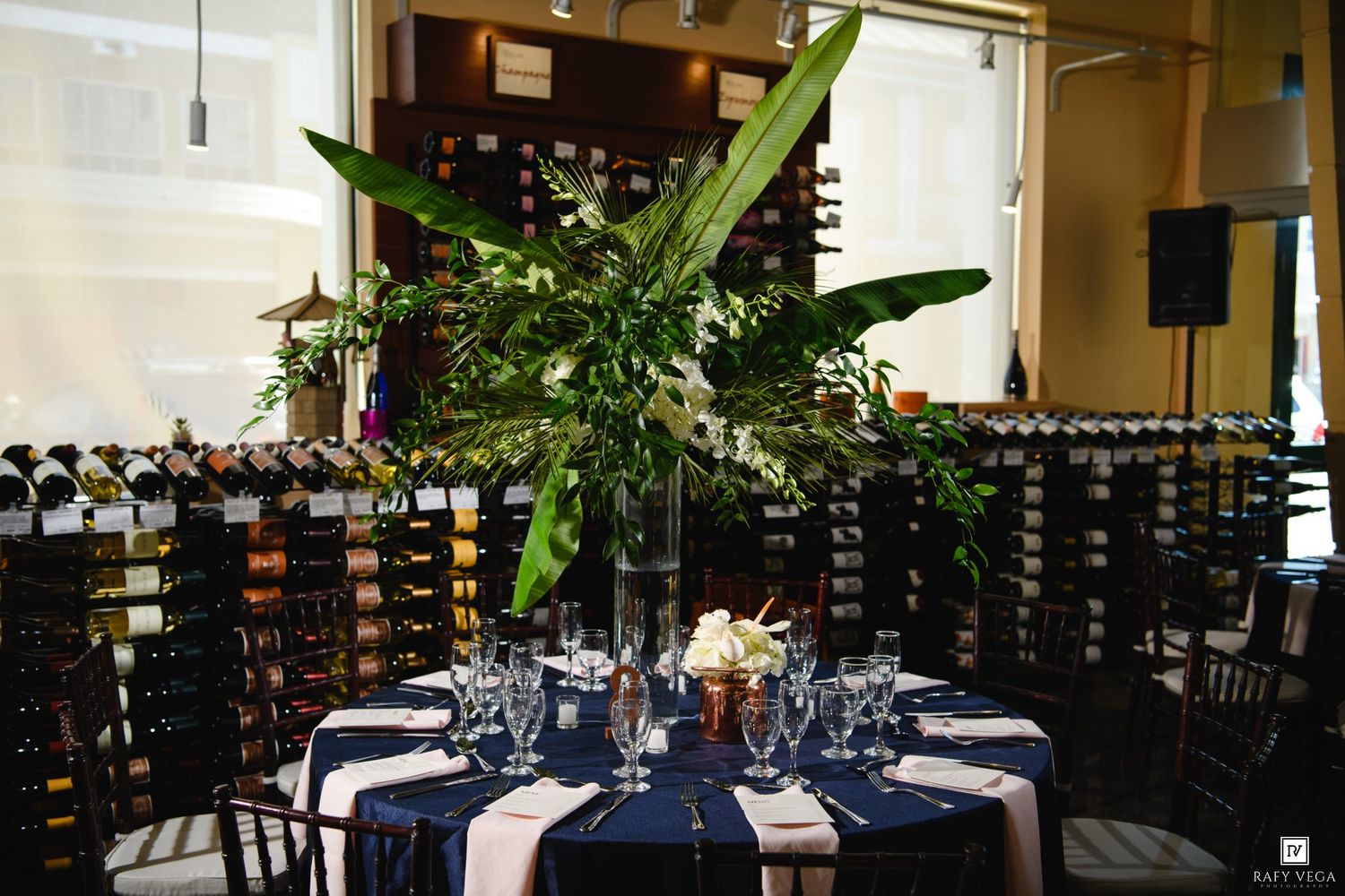 Elegant wedding reception table with navy linens, tropical floral centerpiece, and wine bottle wall backdrop.
