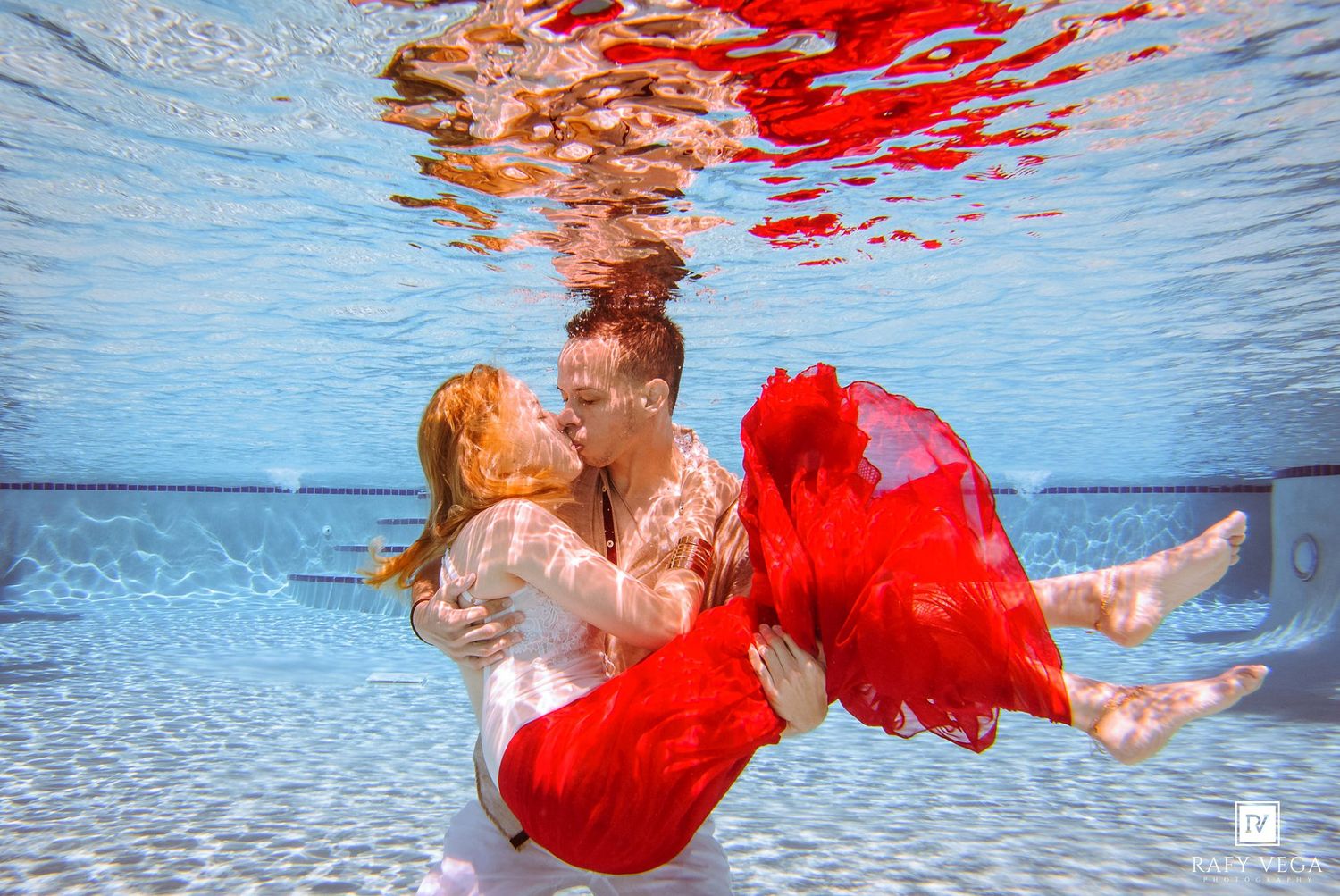 Couple sharing a romantic underwater kiss in a pool, woman wearing a flowing red dress, captured in stunning aquatic photography.