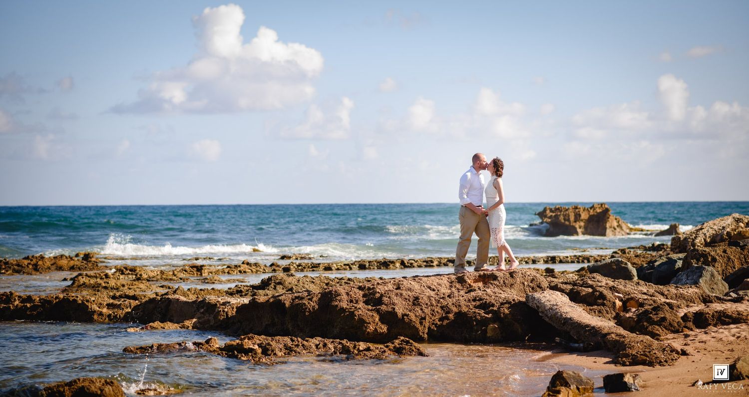 Couple embracing on rocky shoreline with ocean waves and blue sky backdrop, ideal for beach wedding photography.
