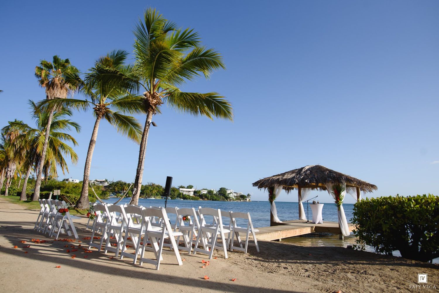 Beachside wedding ceremony setup with white chairs, palm trees, and a thatched gazebo overlooking calm blue waters.