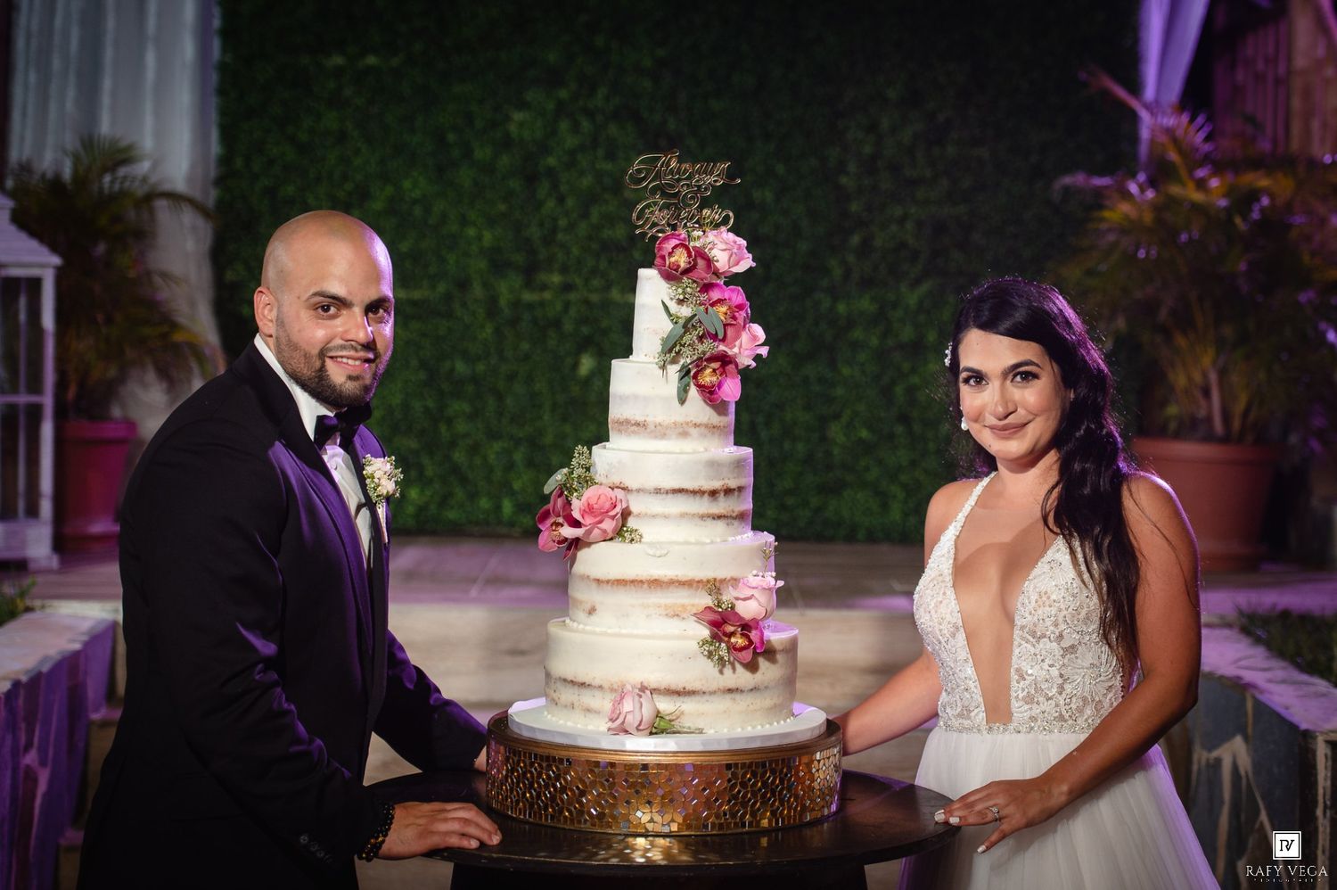 Bride and groom smiling beside a elegant multi-tier white wedding cake adorned with pink flowers.