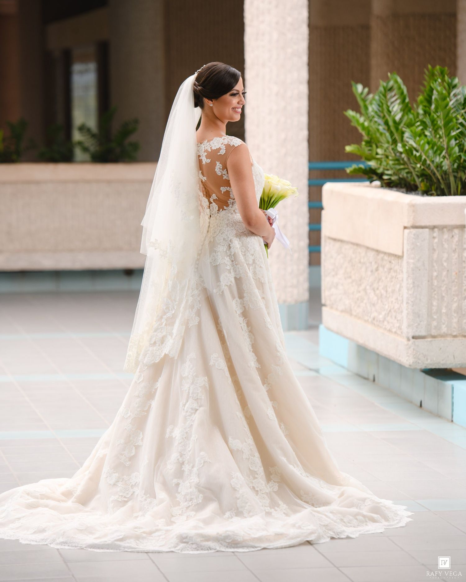 Bride in elegant lace wedding gown with veil and bouquet, showcasing intricate back detail.