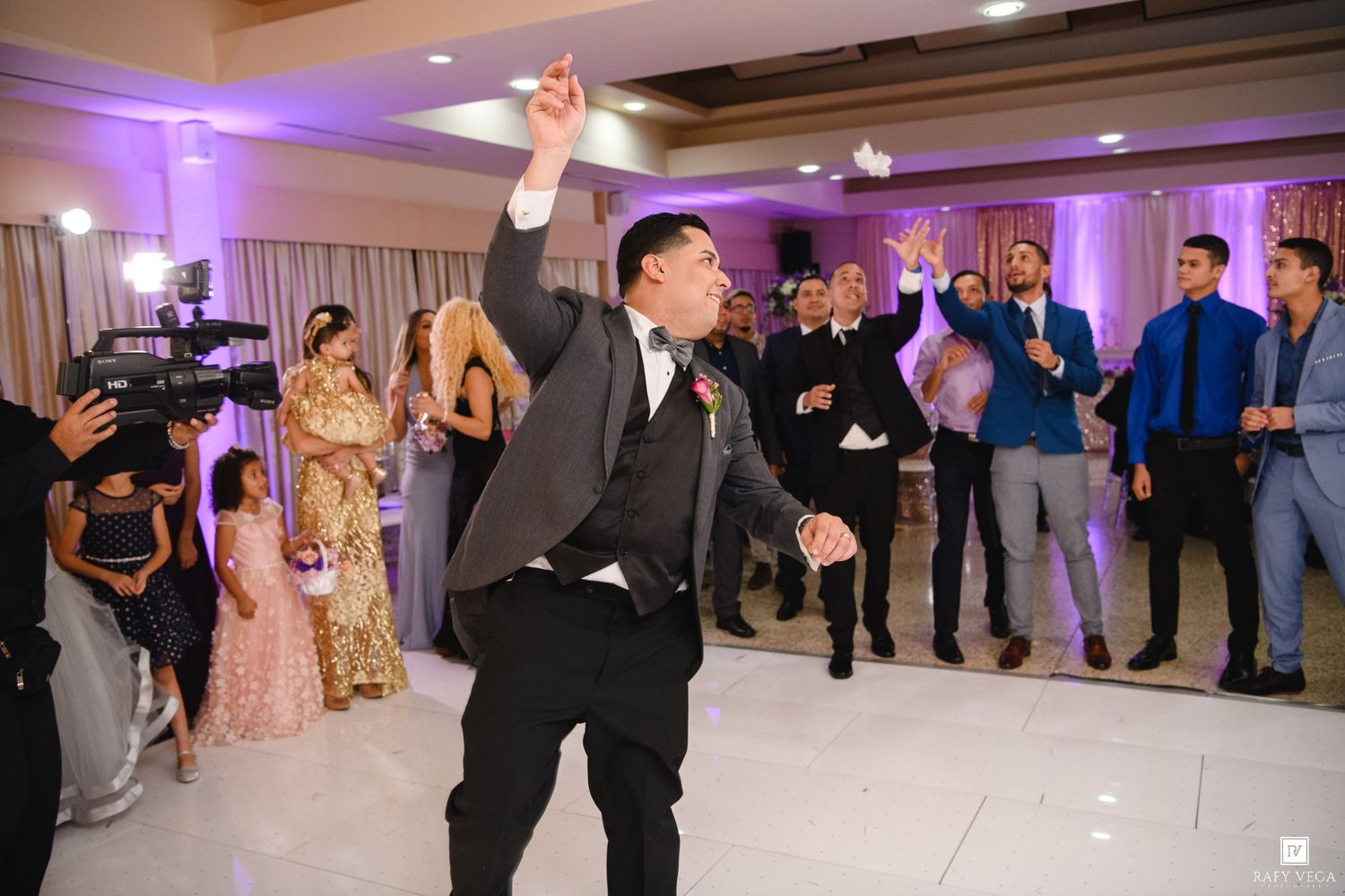 Groom dancing energetically at wedding reception while guests watch and celebrate in elegant banquet hall with purple lighting.