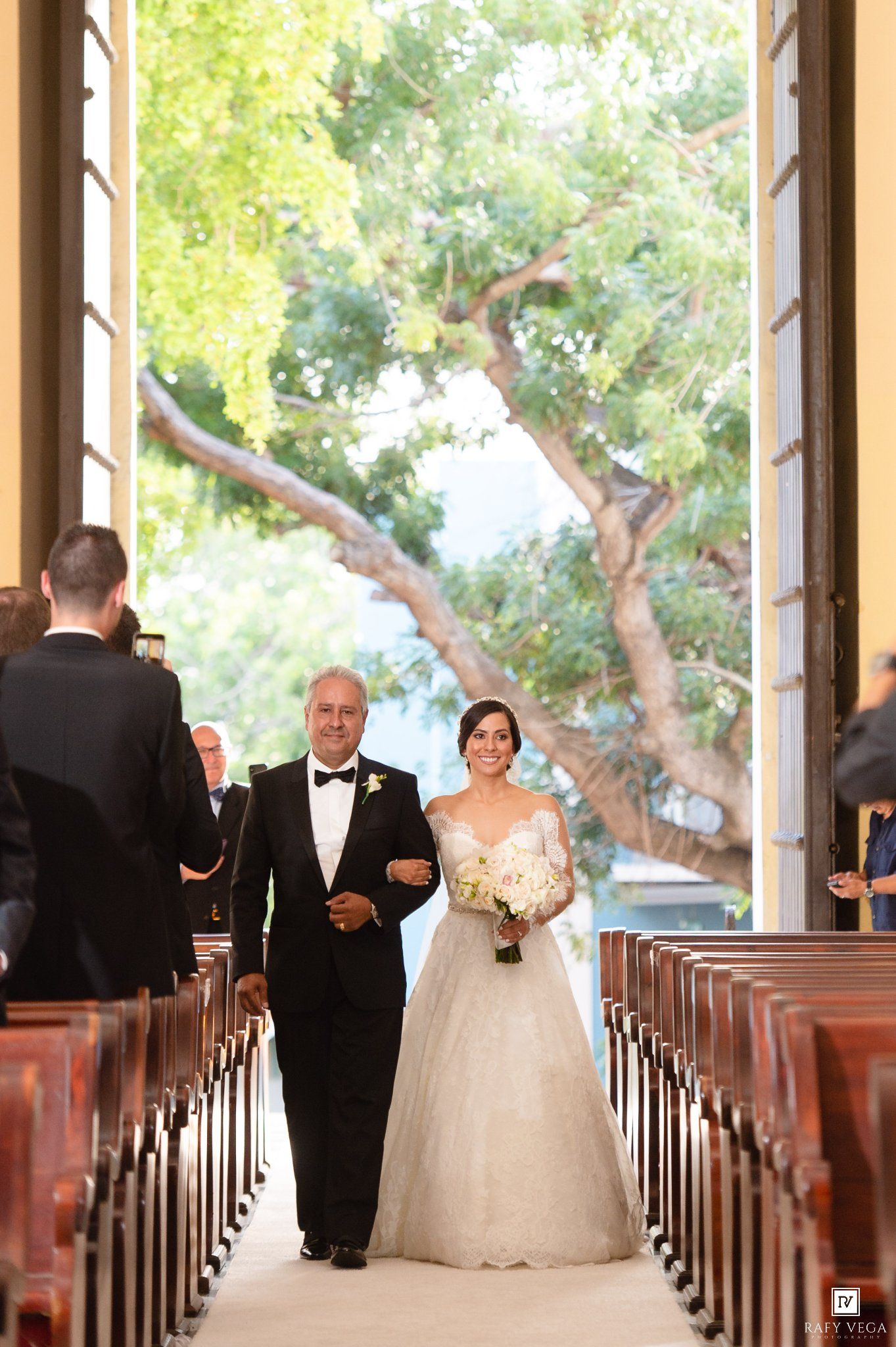 Bride walks down chapel aisle with escort, holding white bouquet, in elegant strapless gown, with sunlit trees visible through open doors.