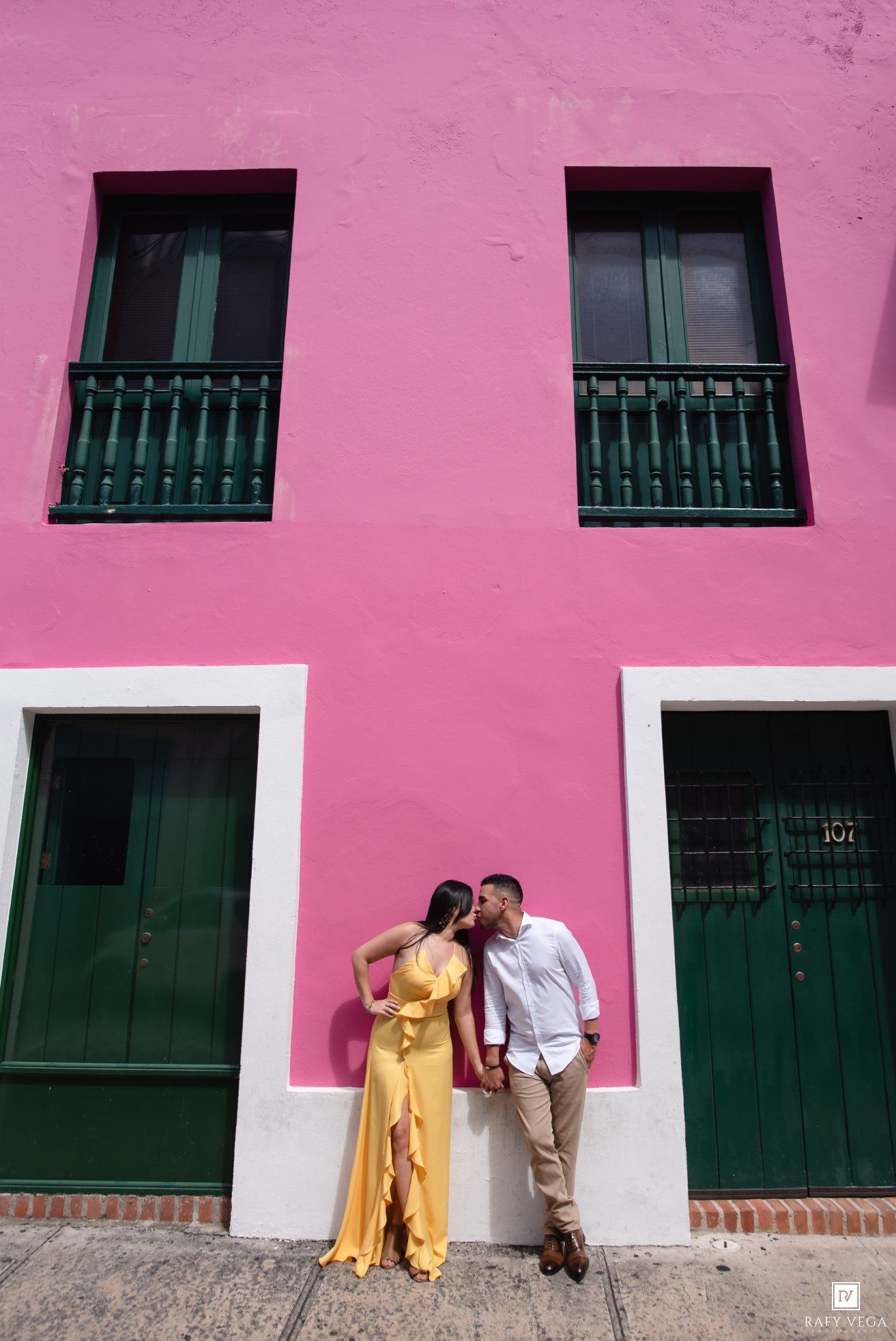 Couple sharing a romantic kiss against a vibrant pink building with green doors and balconies in Old San Juan.
