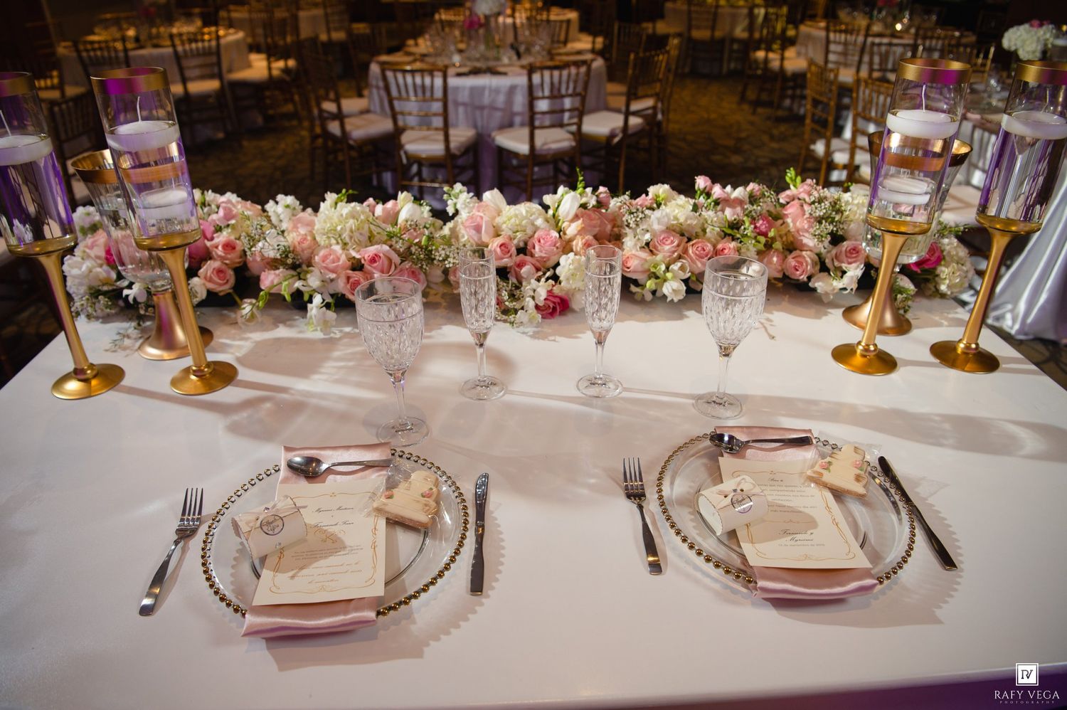 Romantic wedding reception table with pink floral centerpieces, gold candleholders, and crystal glassware on white linen.