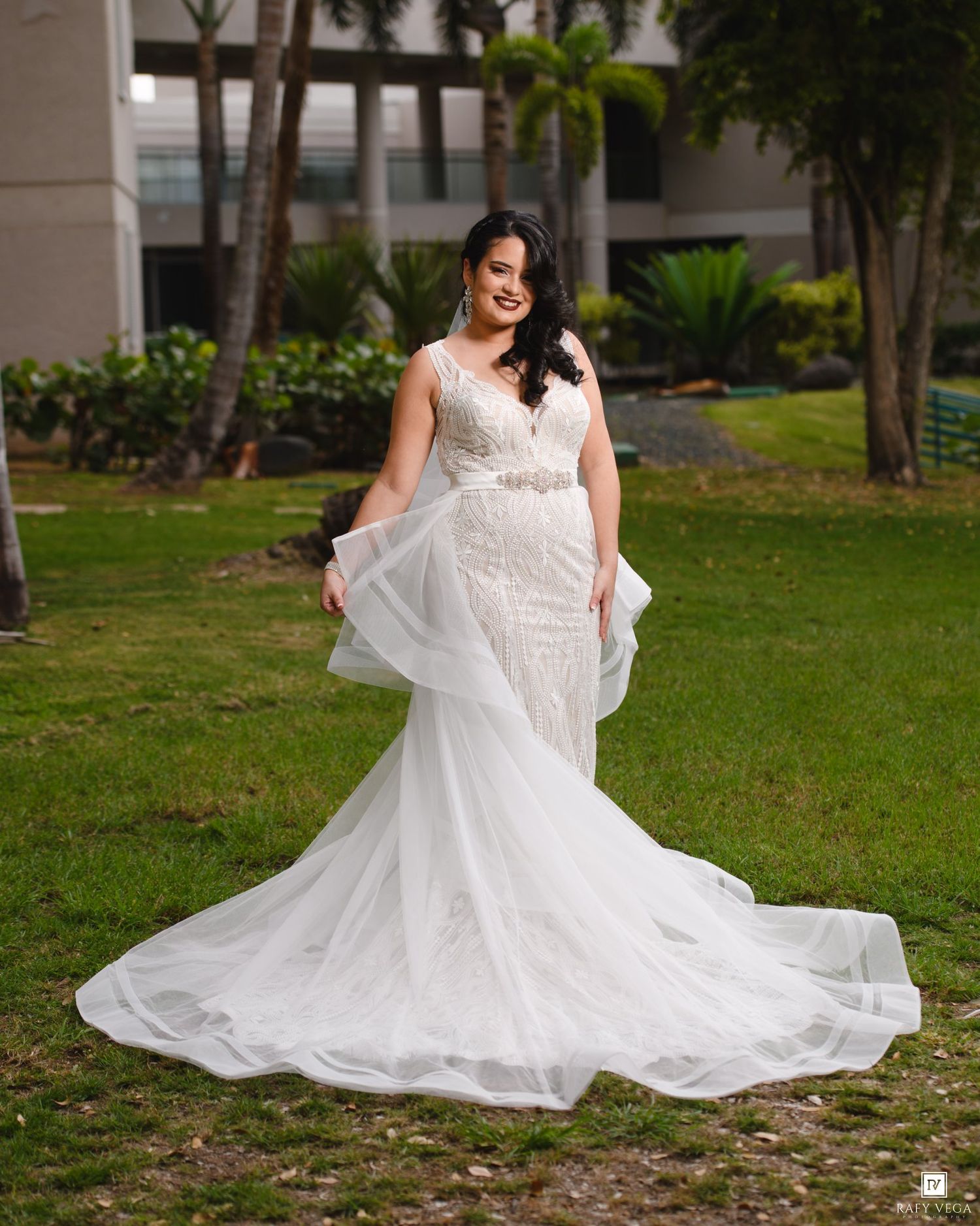 Bride in a white lace mermaid wedding gown with a long train poses outdoors on green grass with palm trees.