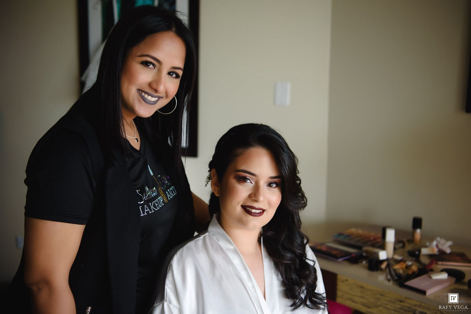 A makeup artist in a black shirt poses with a bride in a white robe during wedding preparations.