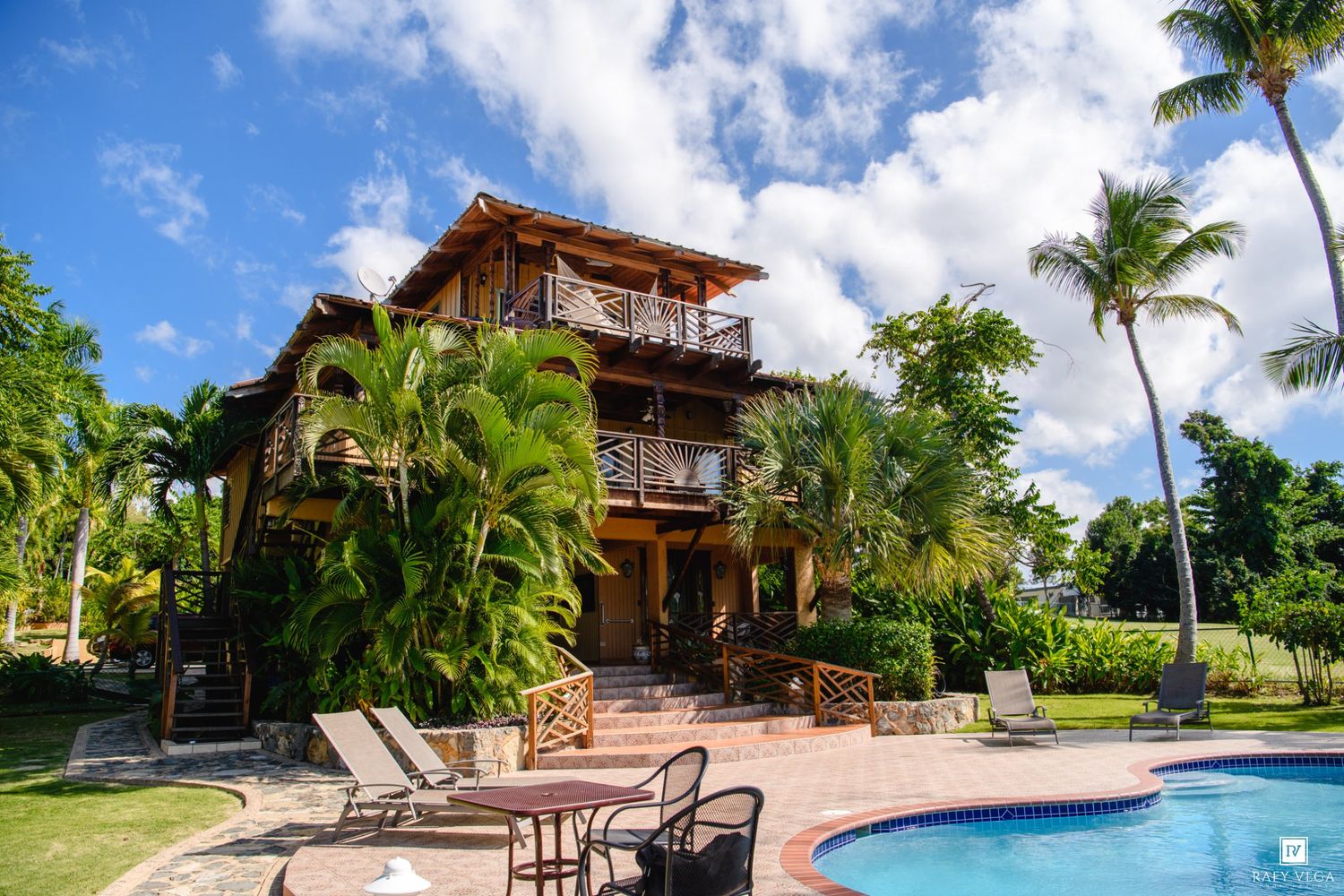 Tropical villa with pool, palm trees, wooden balconies, and lounge chairs under a bright blue sky.