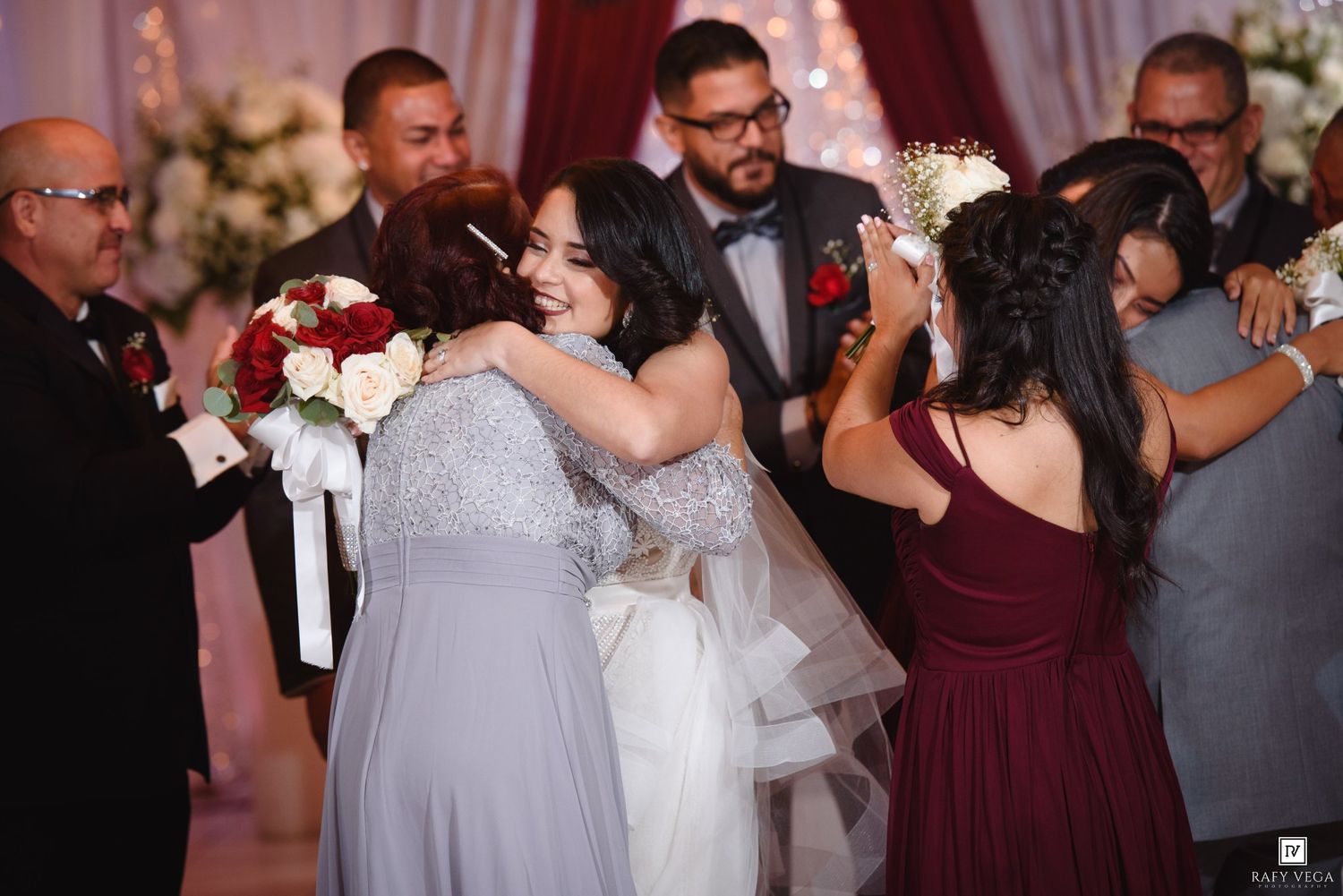 Bride embracing bridesmaid in gray dress during emotional wedding ceremony with guests surrounding them.