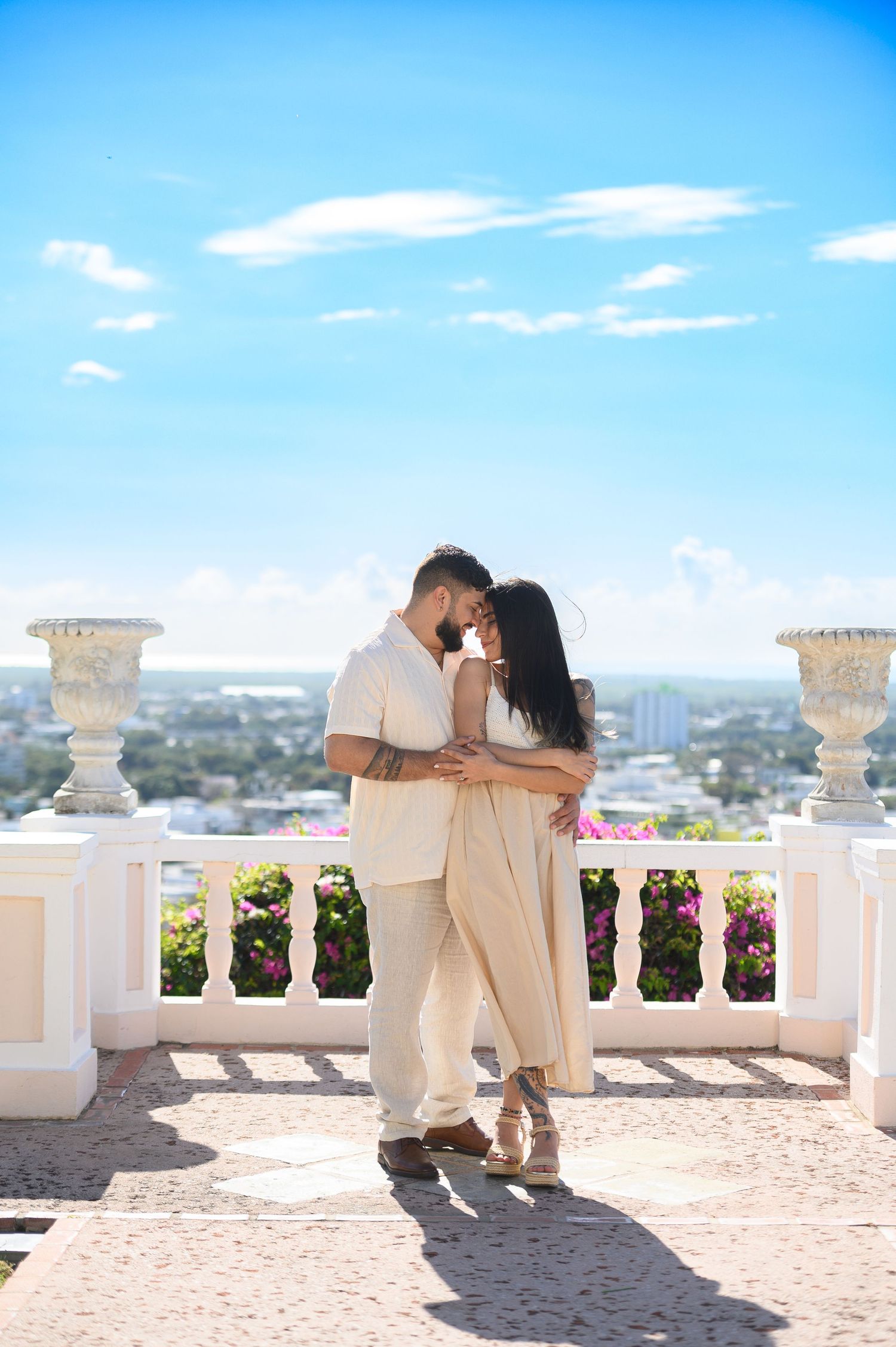 Couple in beige outfits embracing on a white balcony with colorful flowers and blue sky backdrop.