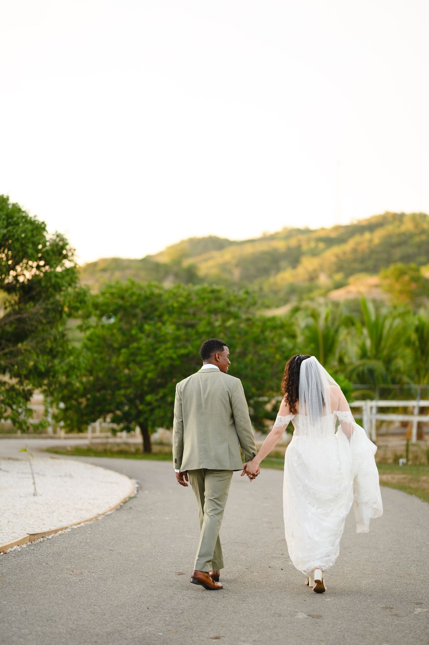 Bride and groom walking hand in hand down a path with lush green hills and trees in the background.
