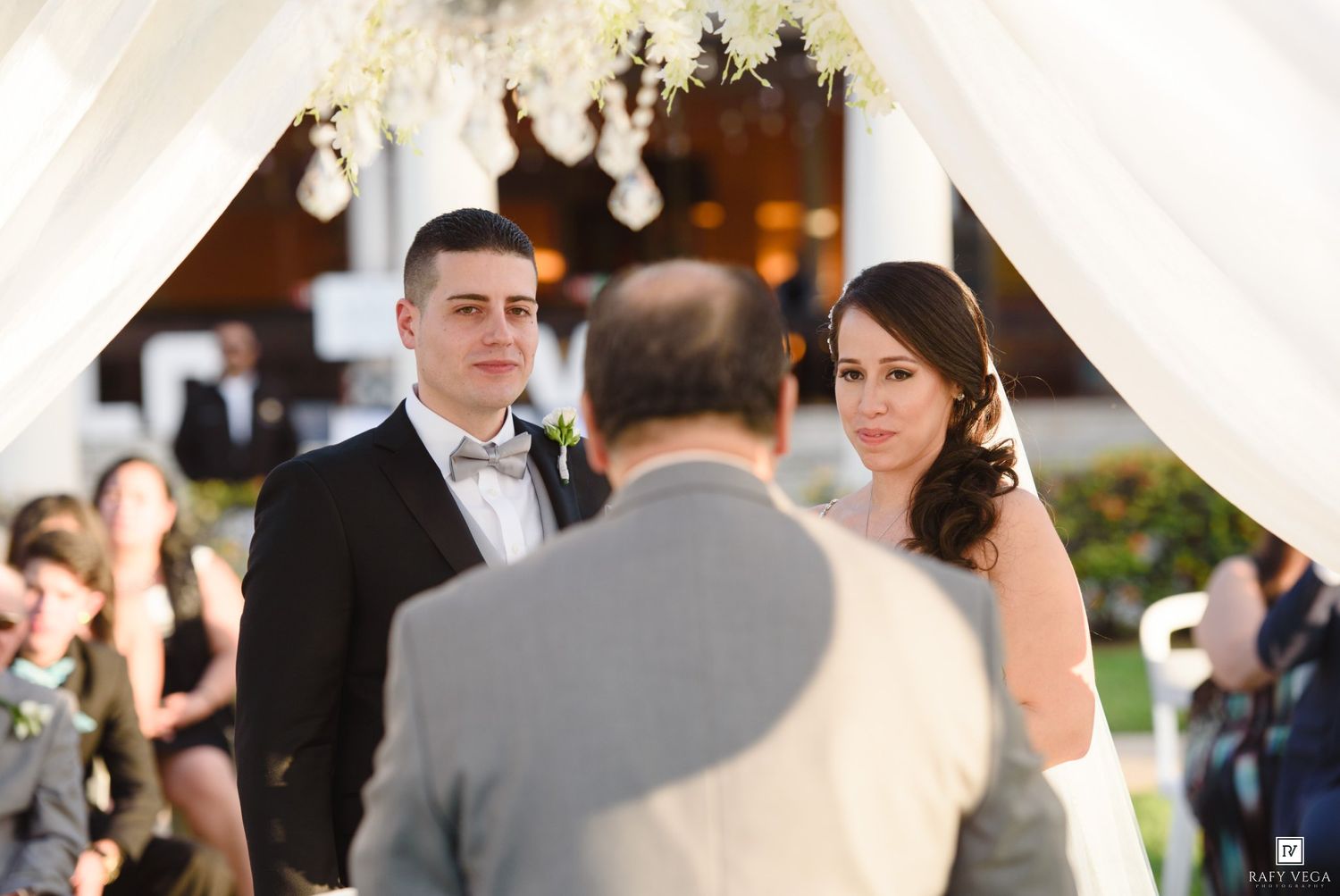 Bride and groom exchange vows under elegant white draped wedding chuppah with floral decorations outdoors.