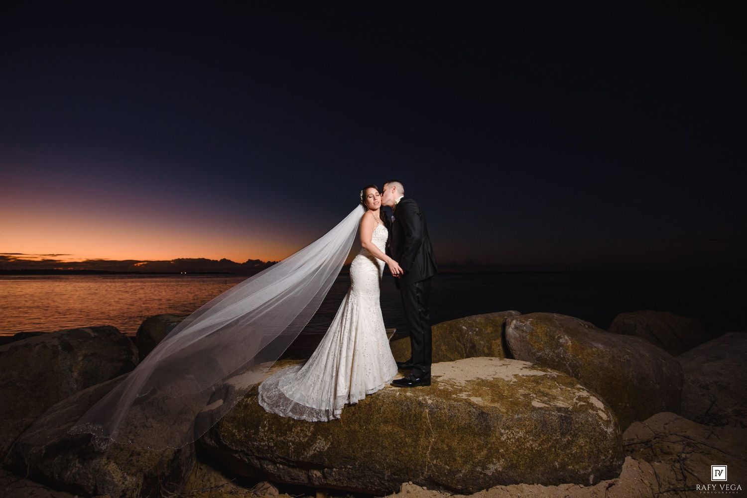Bride and groom share a kiss on rocky shoreline at sunset, with veil flowing in the breeze against a twilight sky.